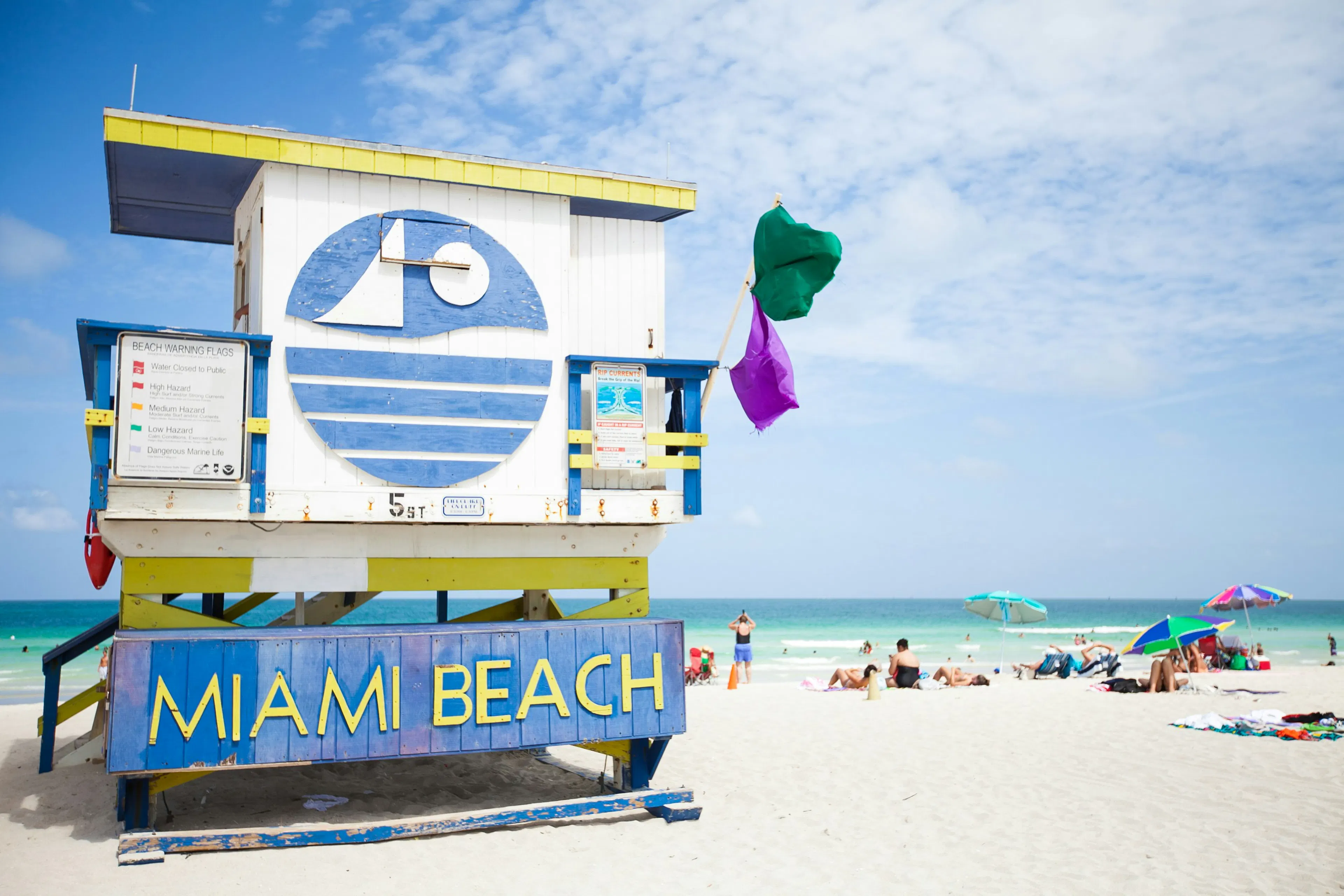 Miami Beach lifeguard tower beside a sandy beach with umbrellas, visitors, and ocean in the background.