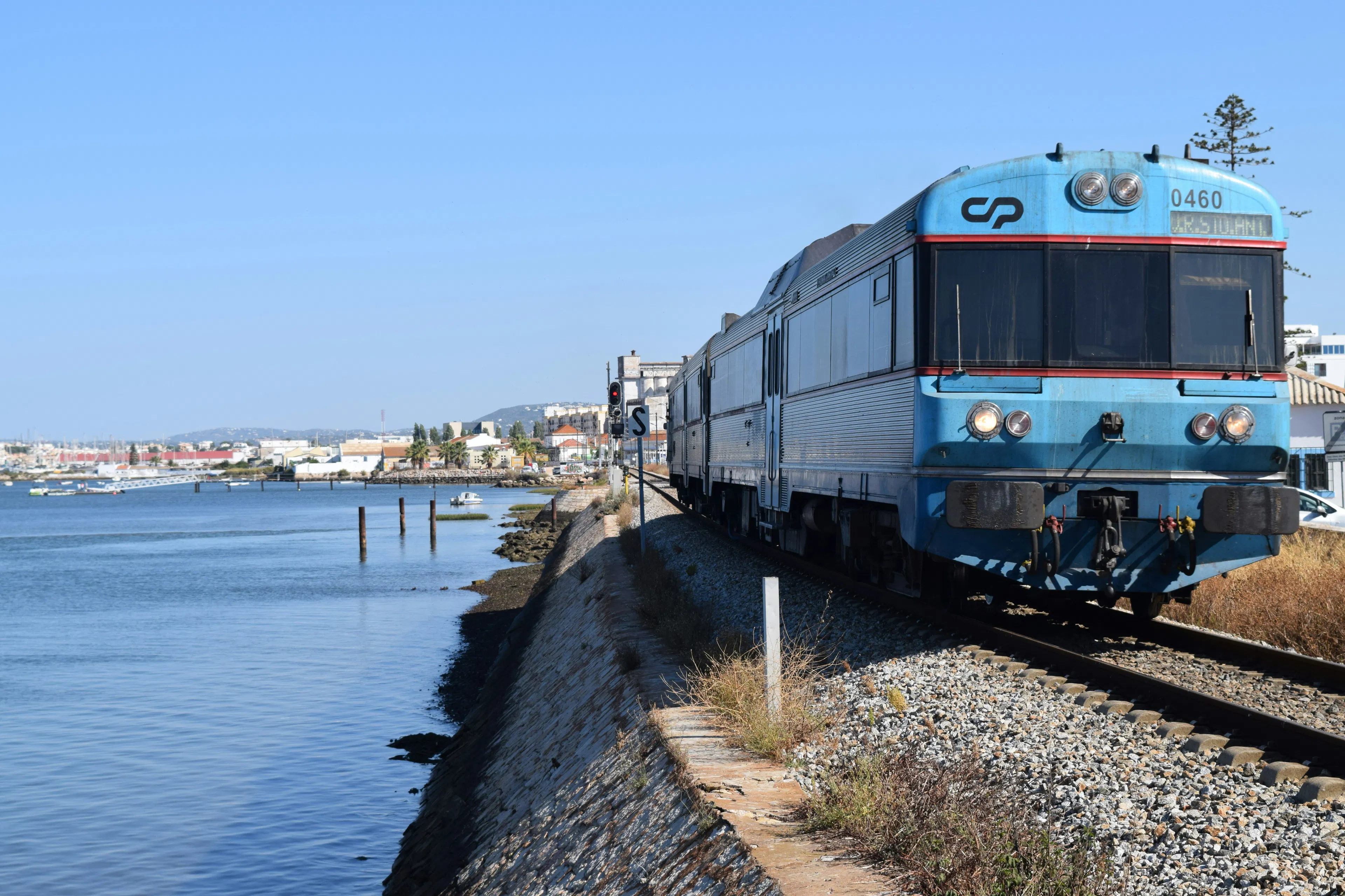 Train traveling along the Faro waterfront, with calm water, coastal views, and the city skyline in the background.