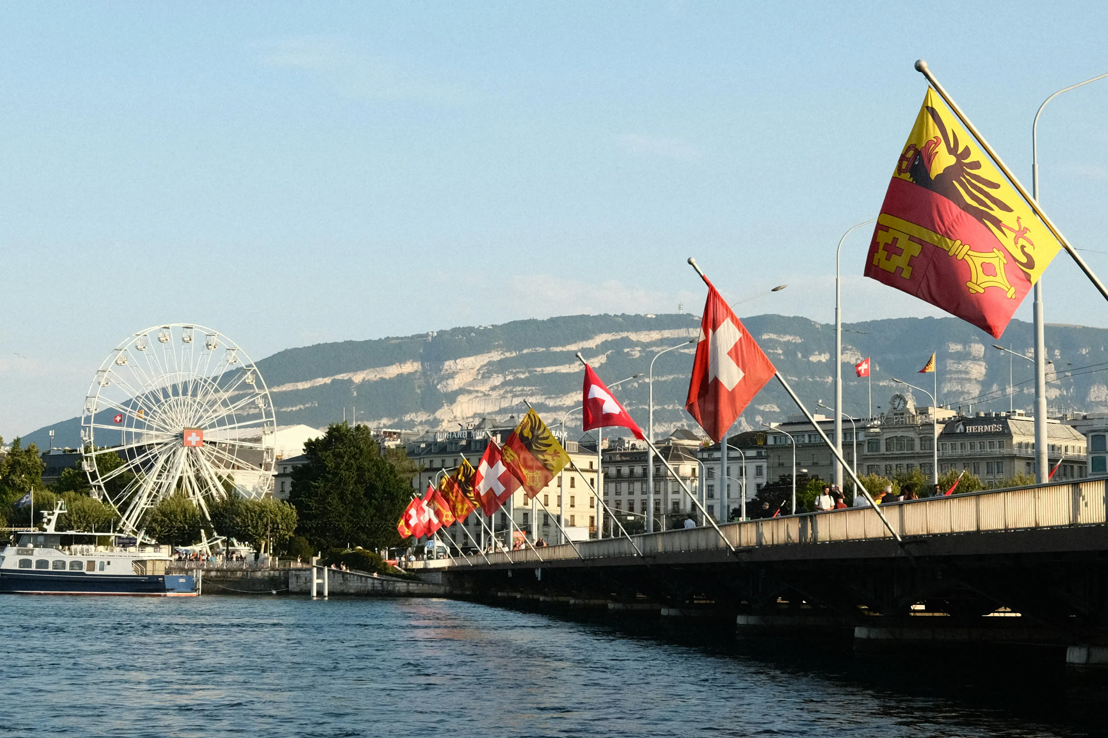Flags of Switzerland and Geneva line a bridge over the Rhone River in Geneva, with a Ferris wheel, waterfront buildings, and distant mountains in the background.