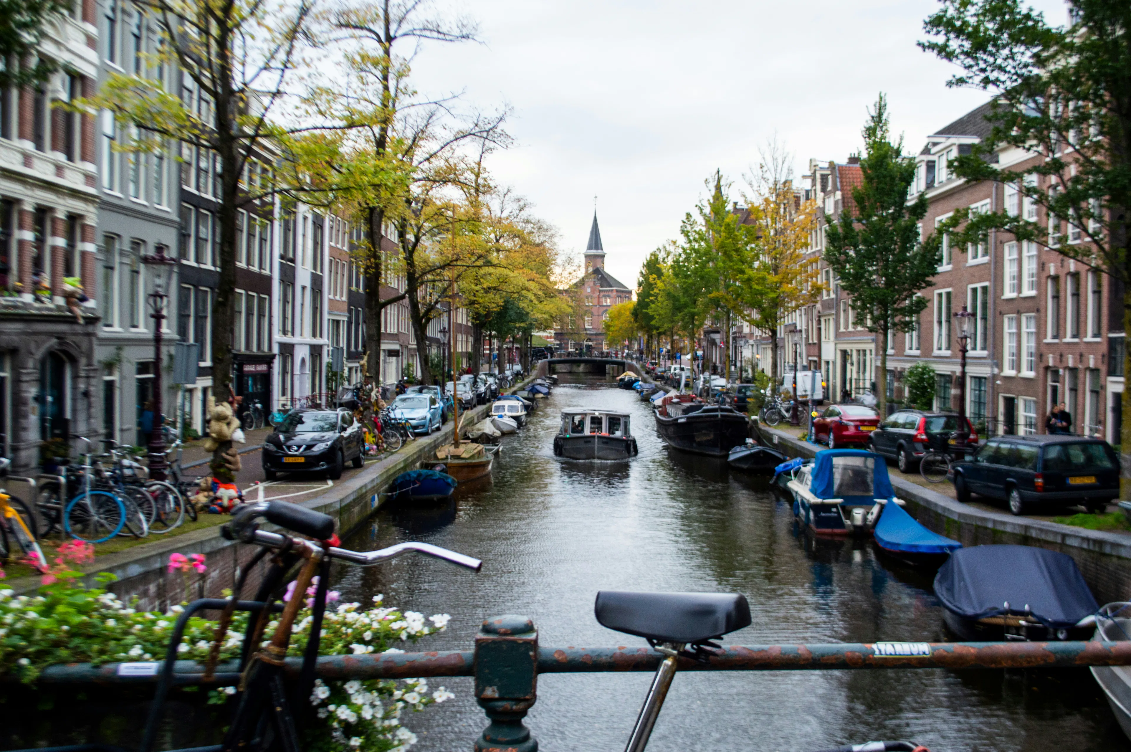 Historic Amsterdam canal lined with traditional Dutch buildings, trees, boats, and bicycles.