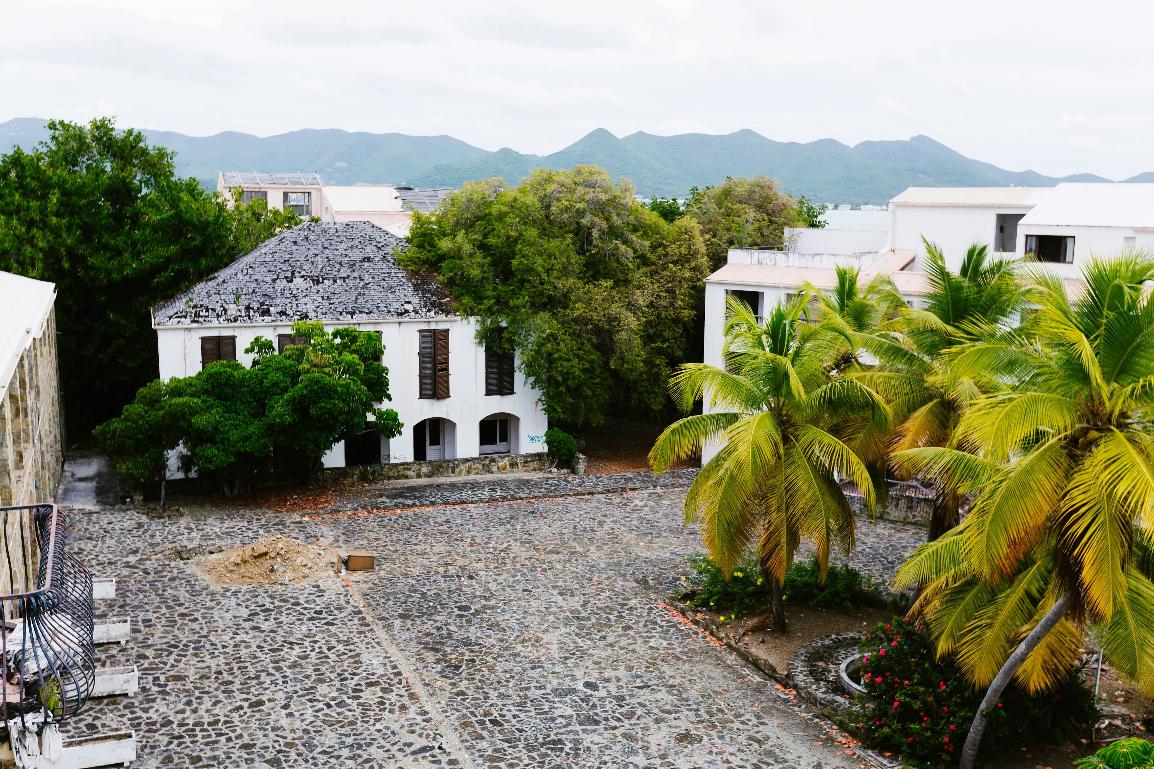 Historic white building surrounded by palm trees and stone pathways with green hills in the background.