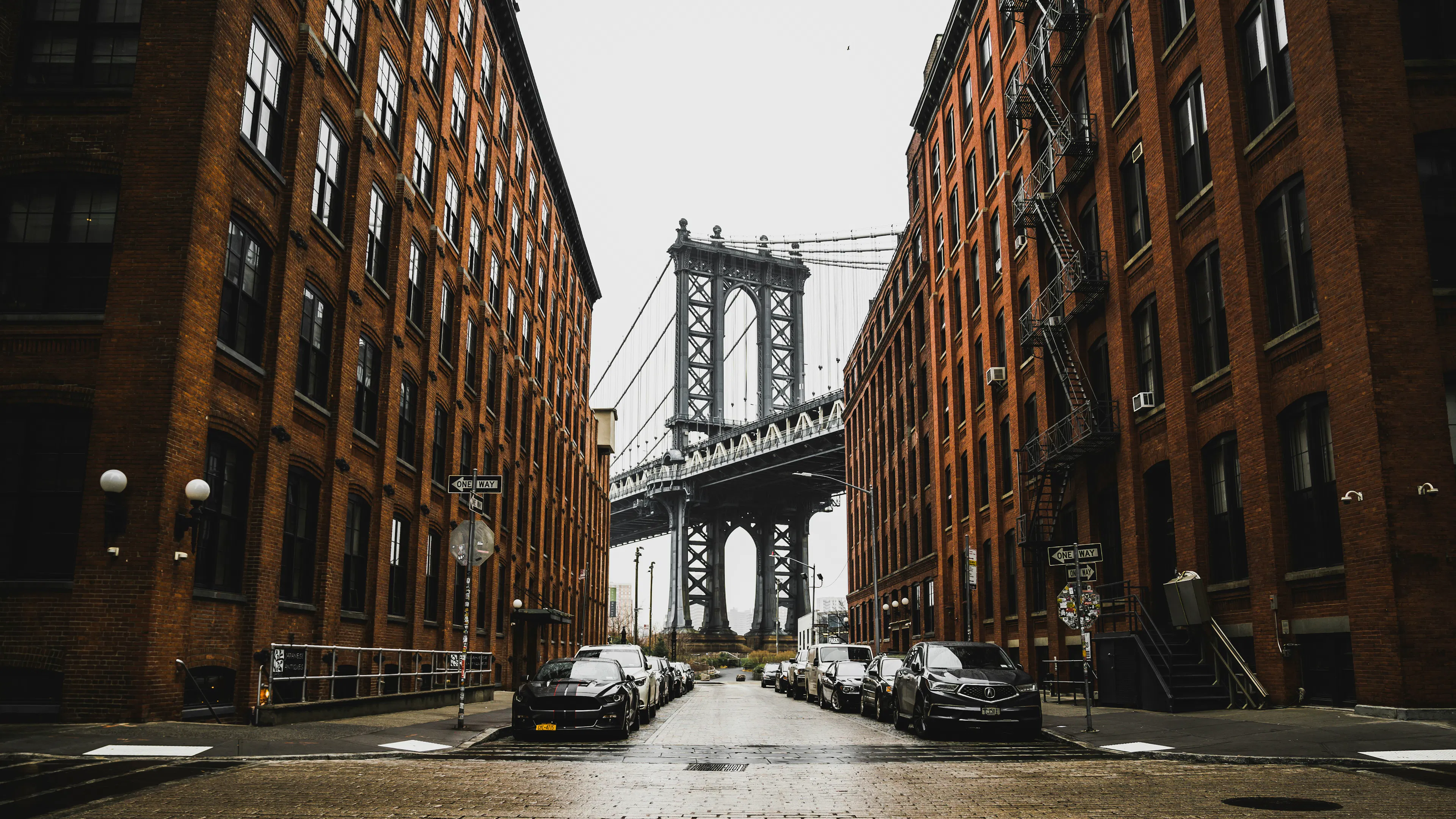 Manhattan Bridge framed by brick buildings and parked cars along a cobblestone street in DUMBO.