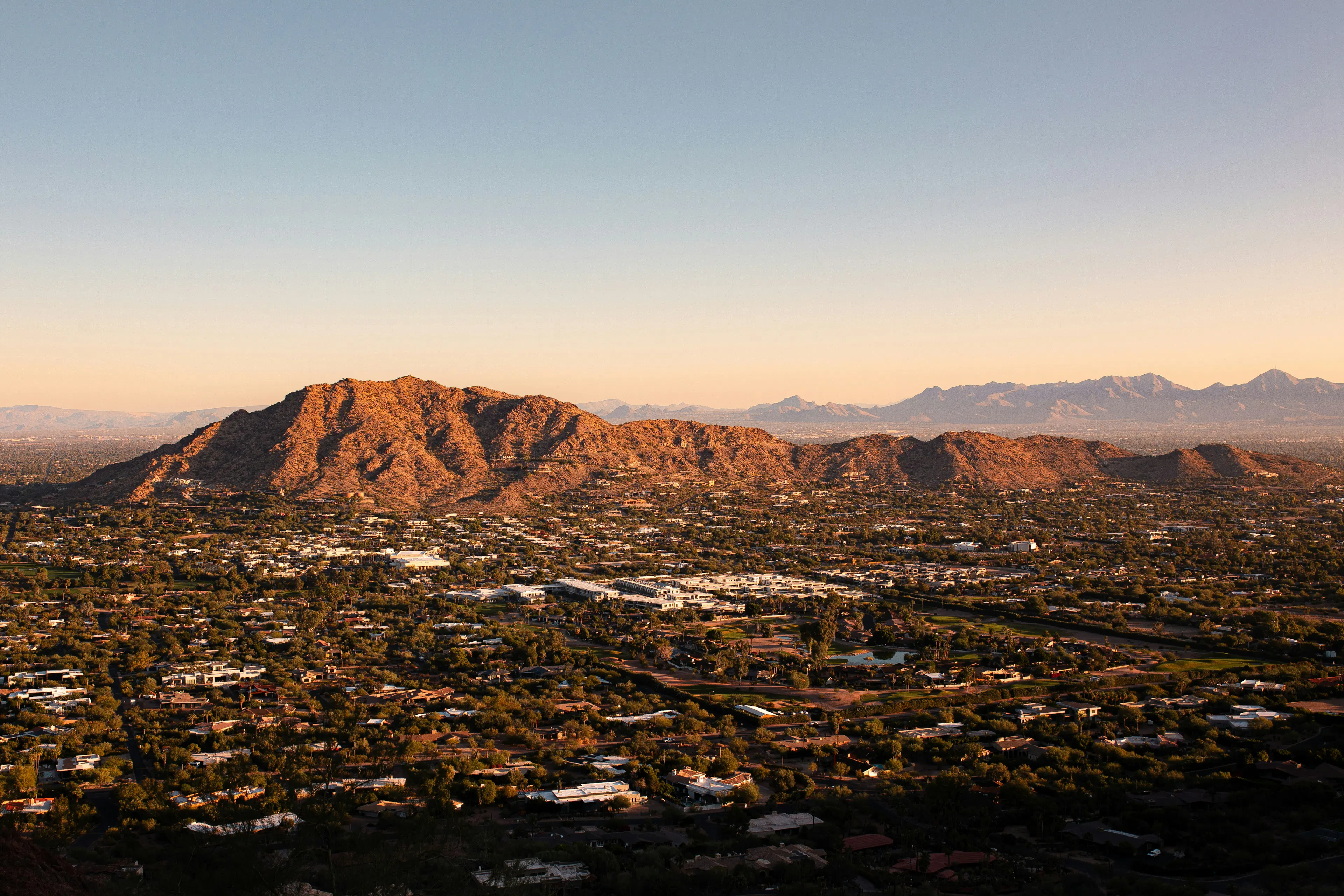 Aerial view of Scottsdale with sunlit desert mountains, neighborhoods, and distant mountain ranges during golden hour.