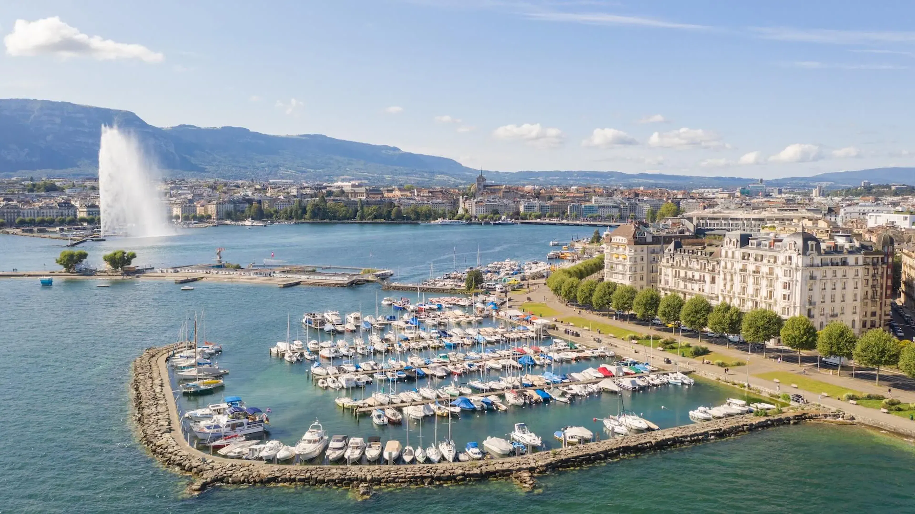 Waterfront cityscape featuring a marina, fountain, and lakeside buildings.