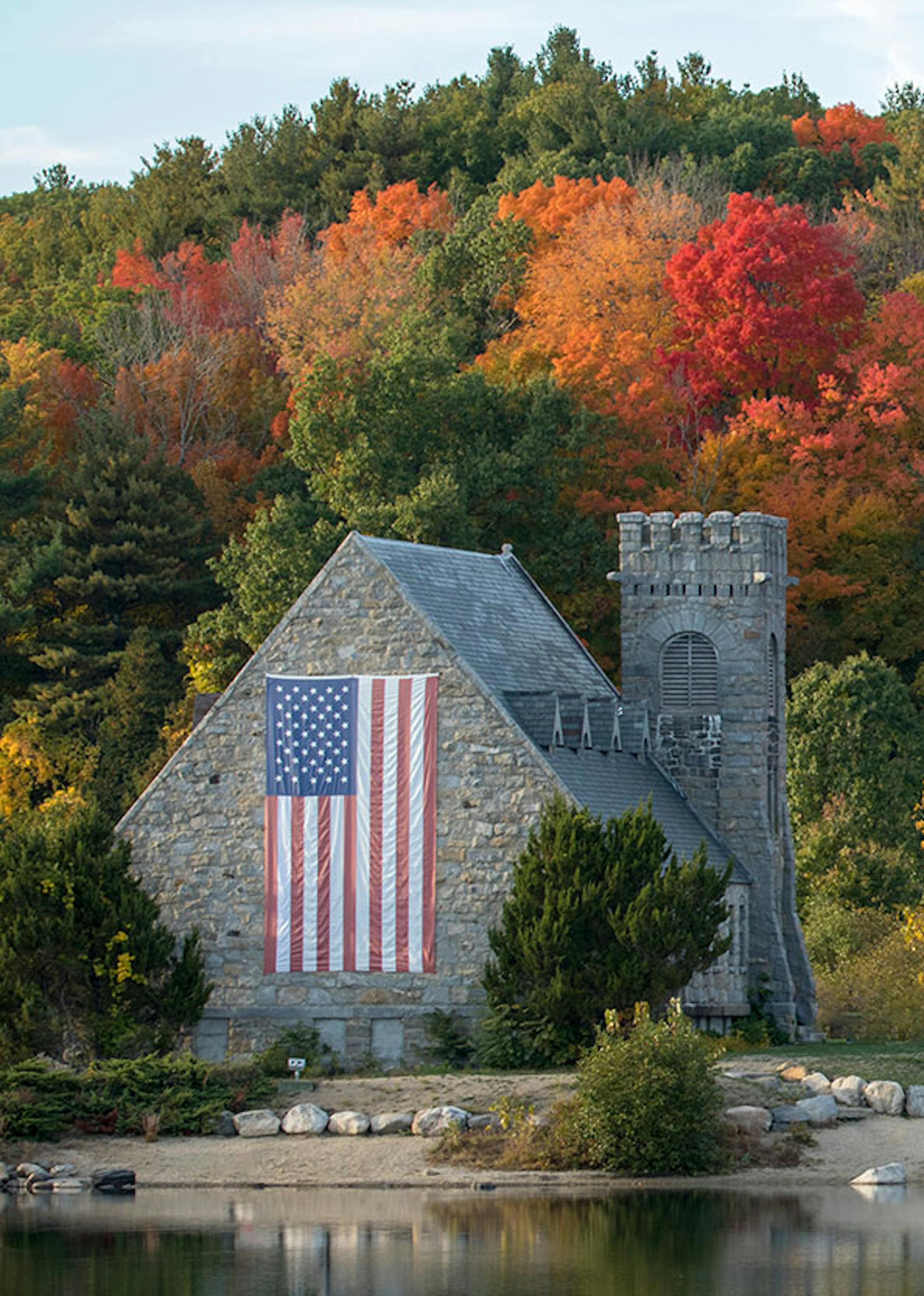 An american flag hangs on the side of a castle structure with fall coloured trees lining a hill surround it