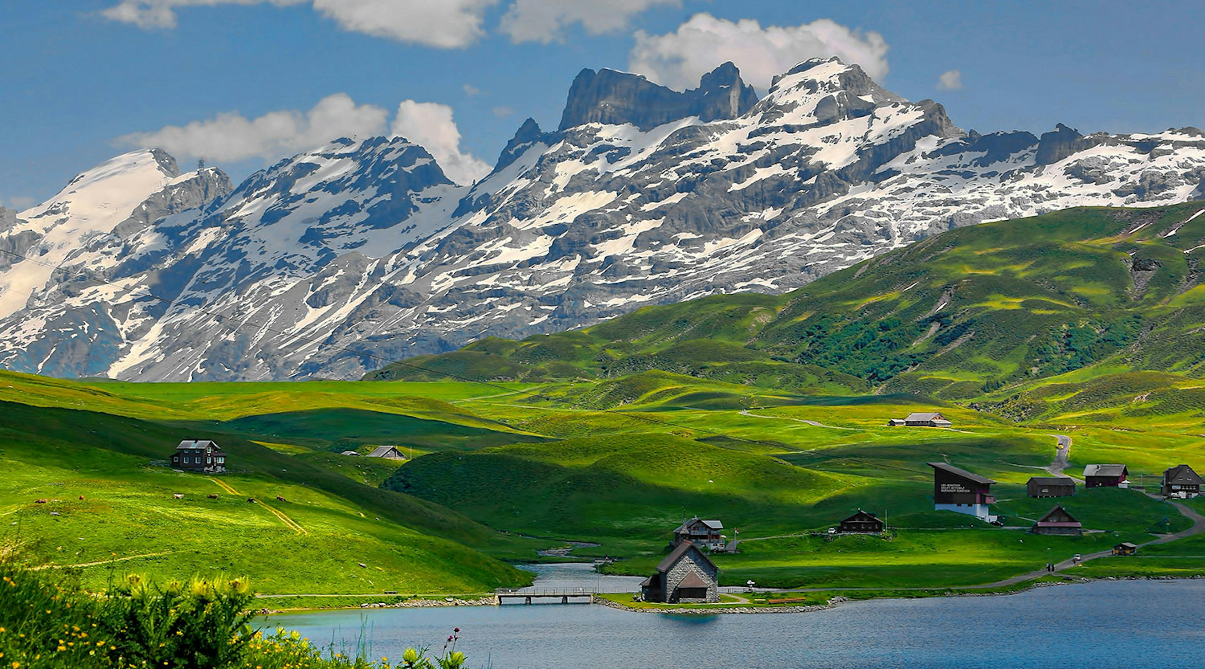 Mountains loom large over a large meadow and a few houses set up against a lake