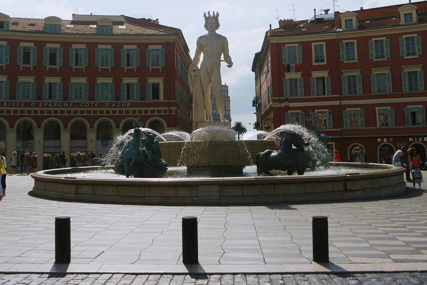 Une statue s'élève, caressée par le jeu d’une fontaine, au cœur d’une place paisible.