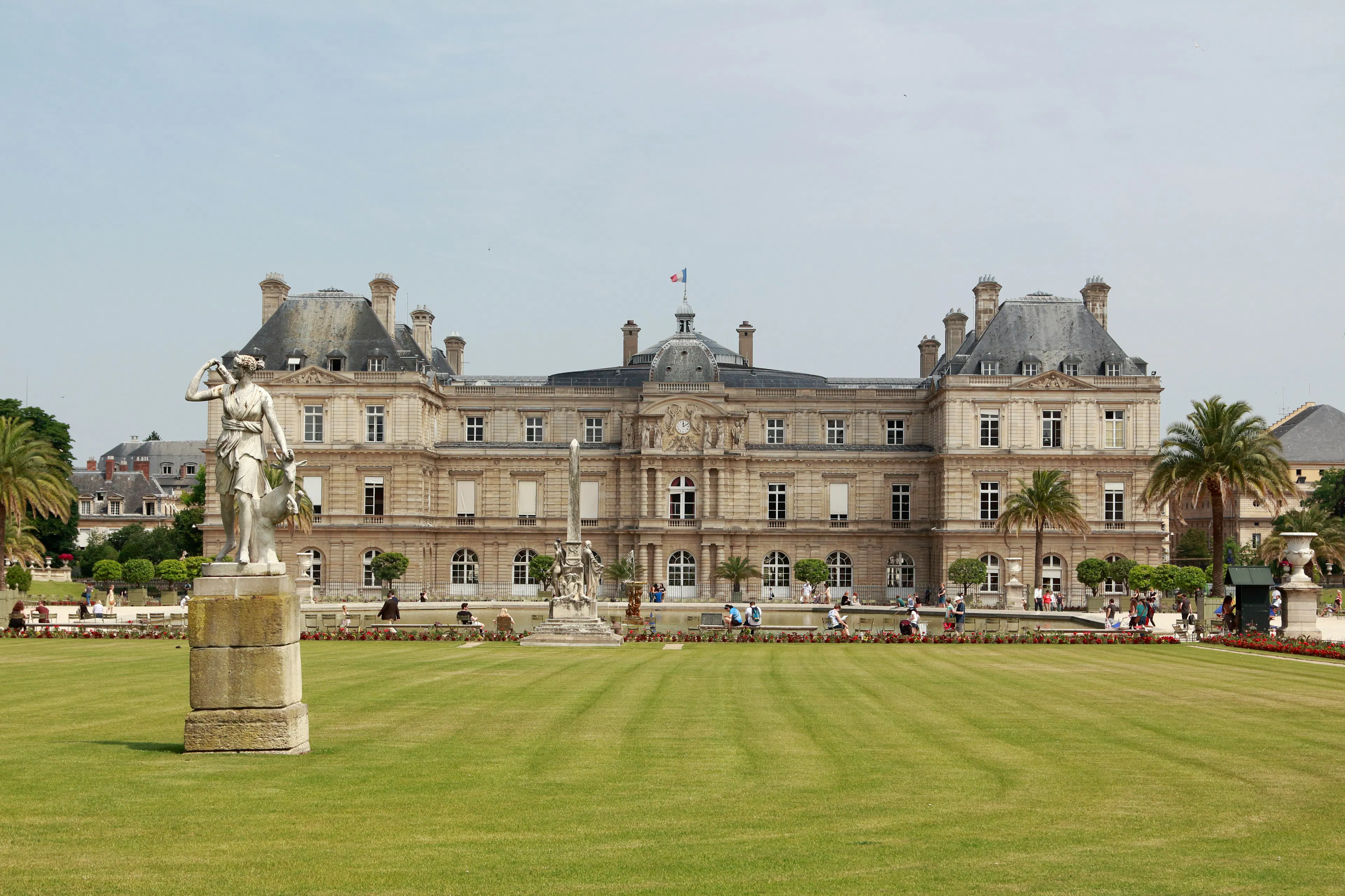 Historic palace in Luxembourg Gardens with statues, palm trees, lawns, and visitors in front.