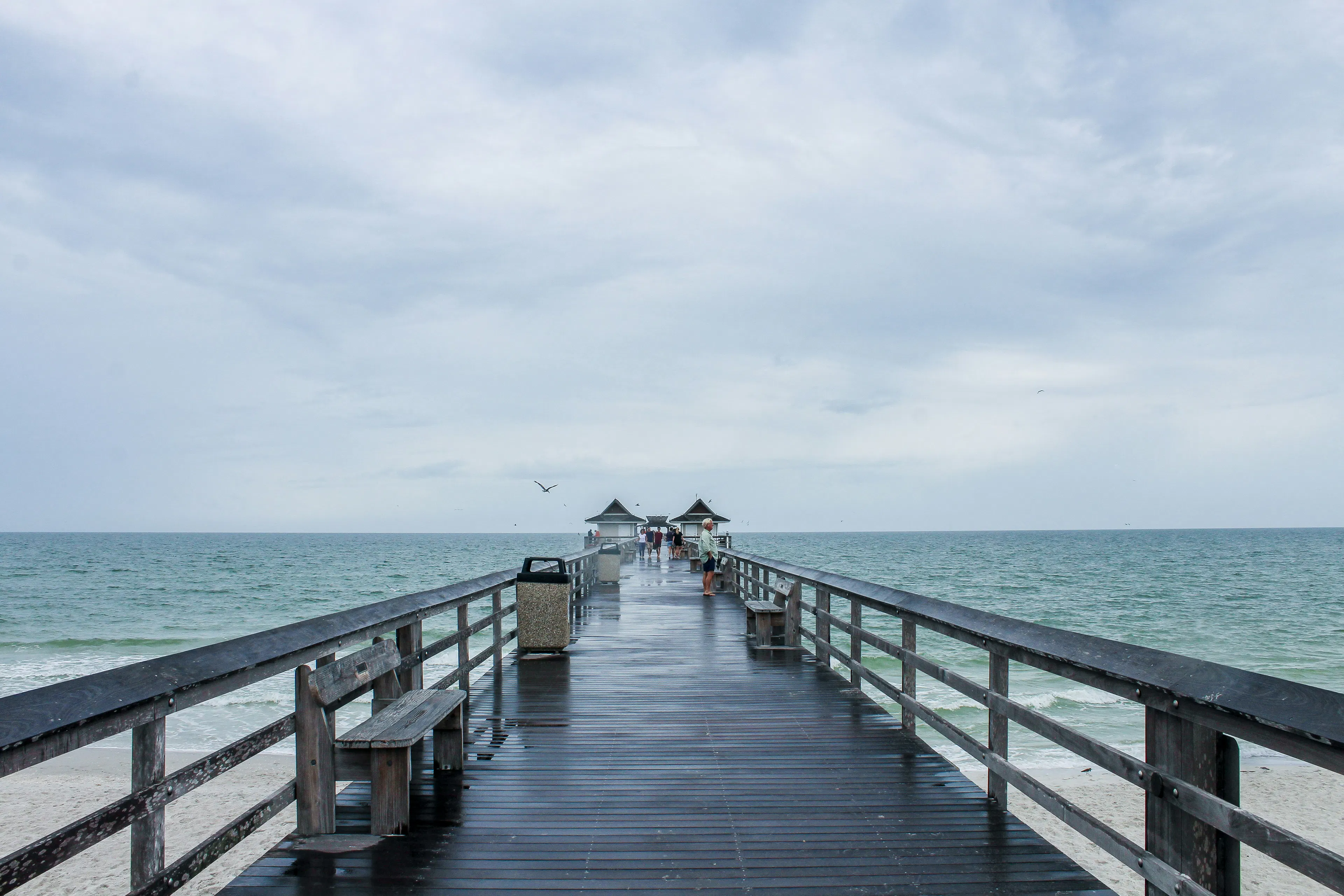 Wooden pier extending into the Gulf of Mexico with benches, visitors, and cloudy skies above calm water.