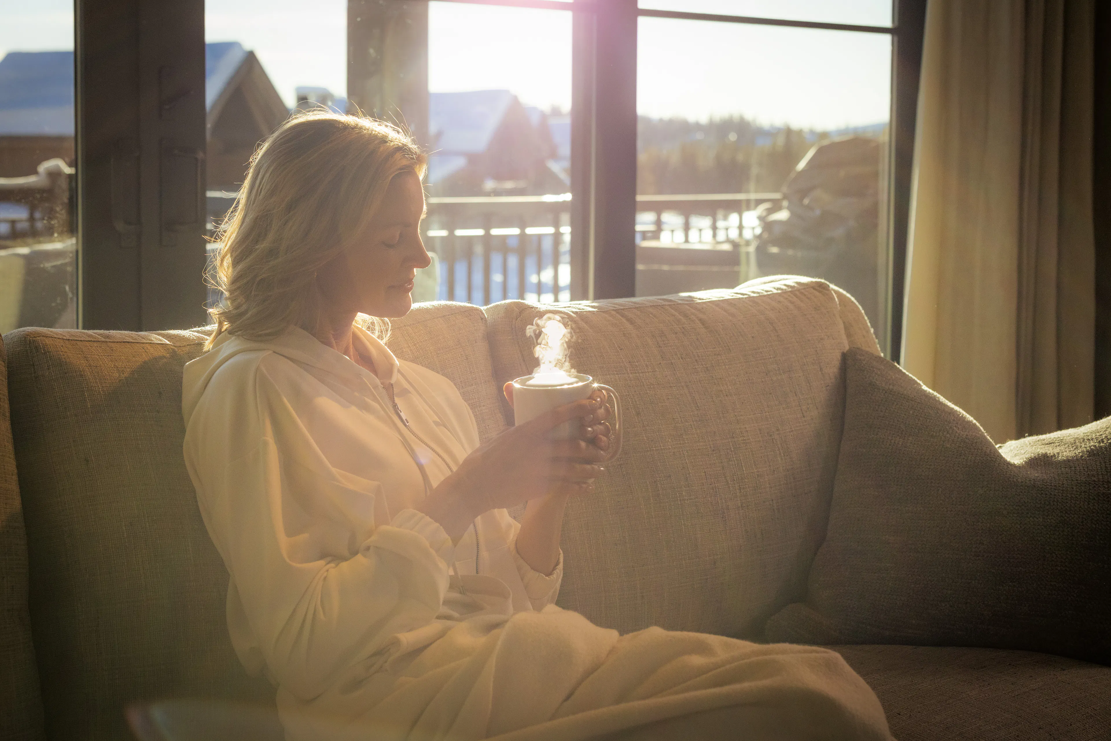 A woman in white sitting on a couch in front of a window with golden sunlight pouring through