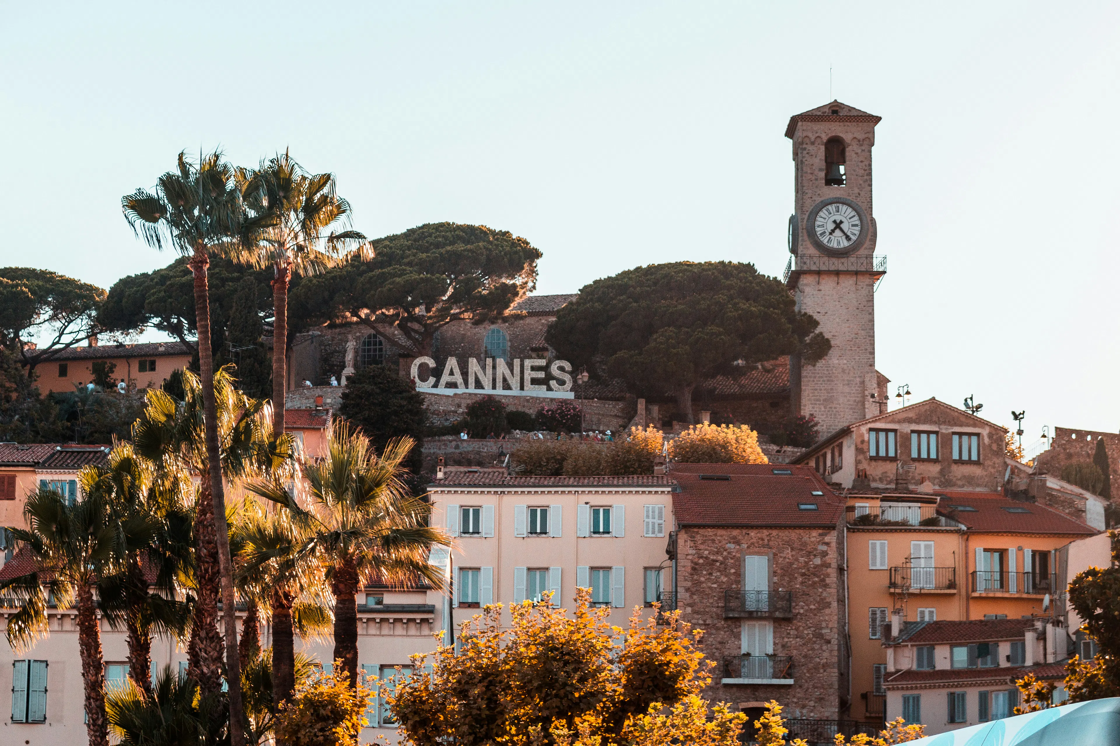 Clock tower above Cannes rooftops with palm trees and hillside buildings in the background.