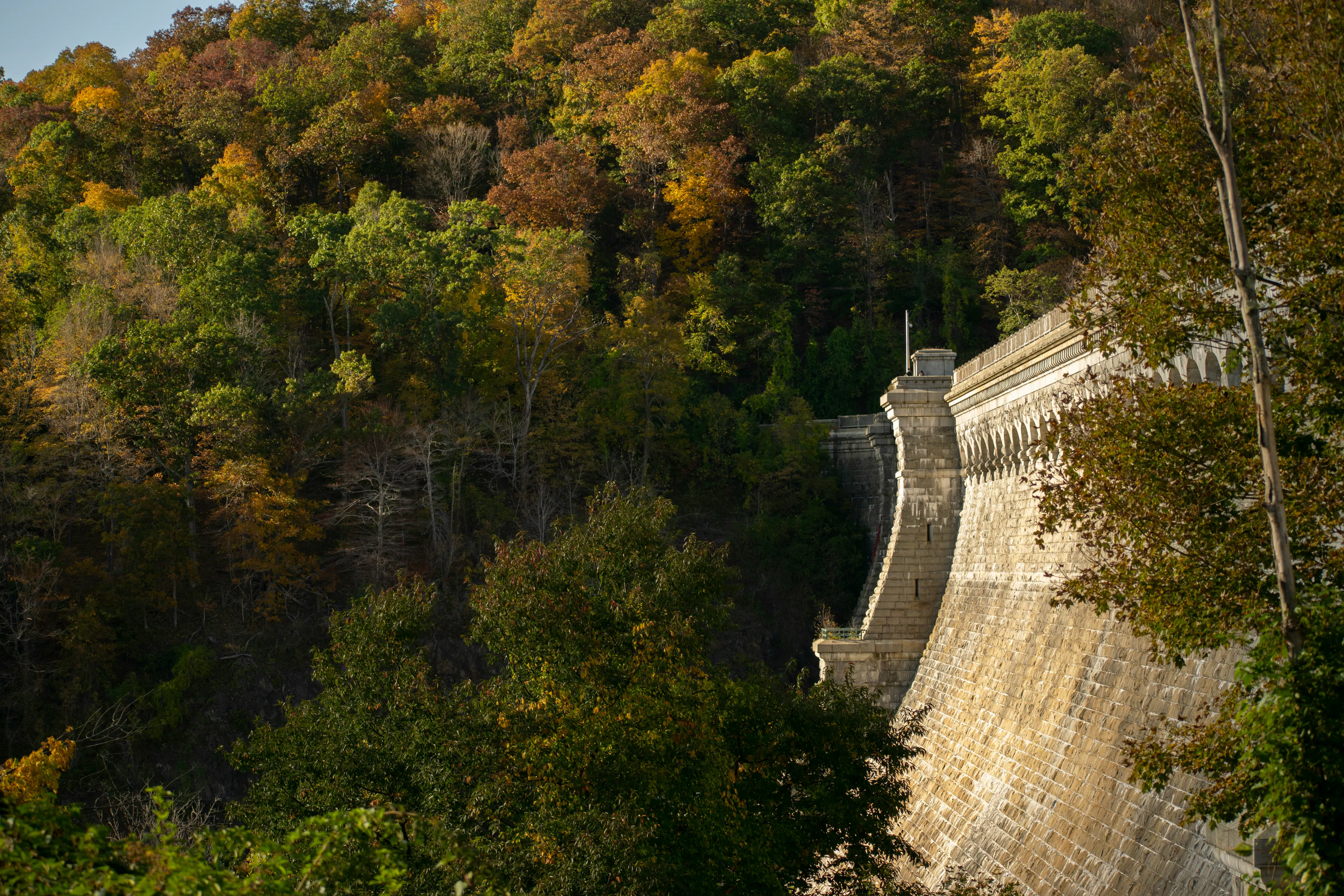 Stone wall of Croton Gorge Dam bordered by colorful autumn trees on a wooded hillside.