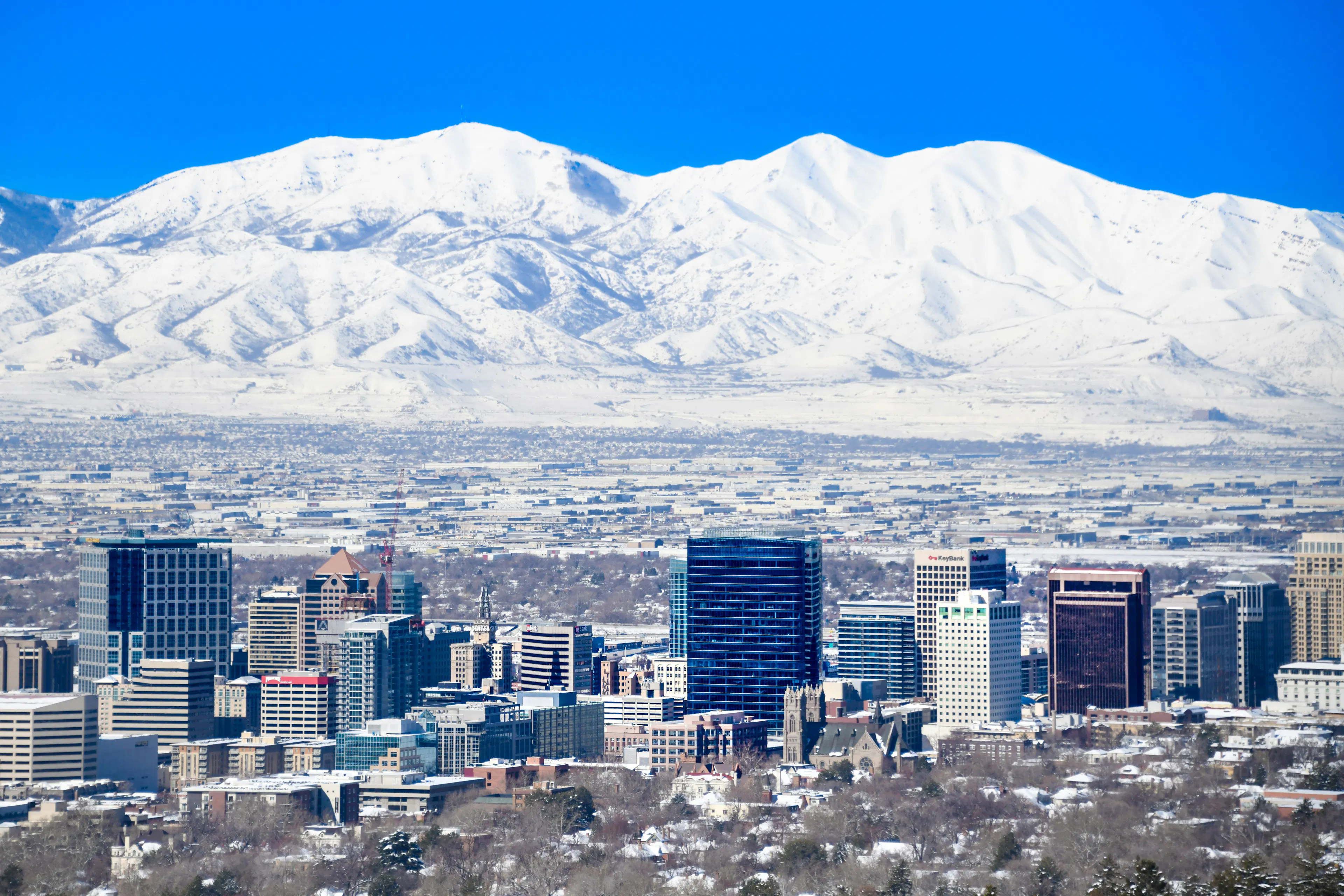 Salt Lake City skyline with snow-covered Wasatch Mountains rising behind downtown buildings under a clear blue sky.