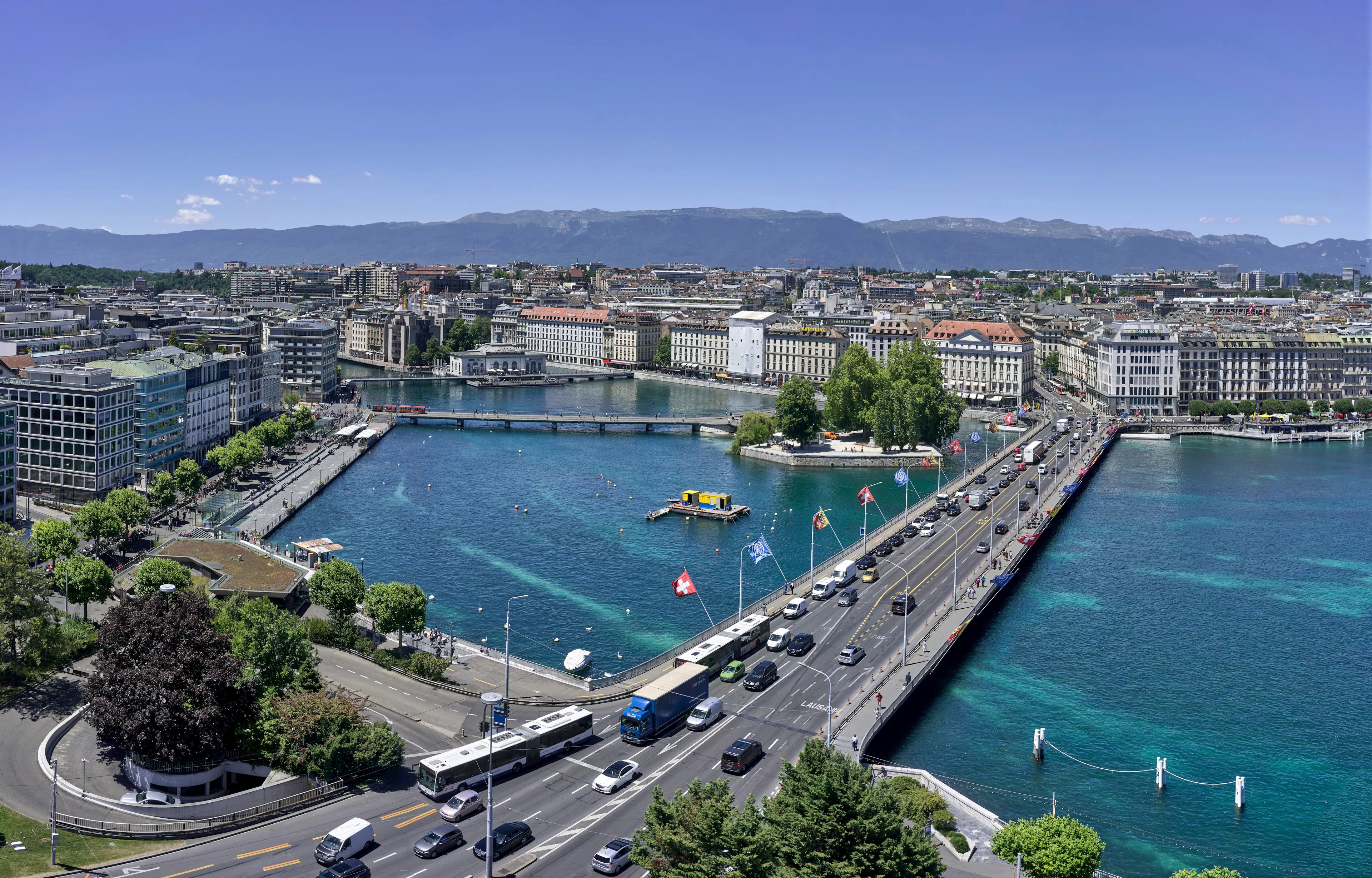 Aerial view of Geneva, Switzerland, showing bridges crossing turquoise waterways, city buildings along the shoreline, and mountains in the distance under a clear blue sky.