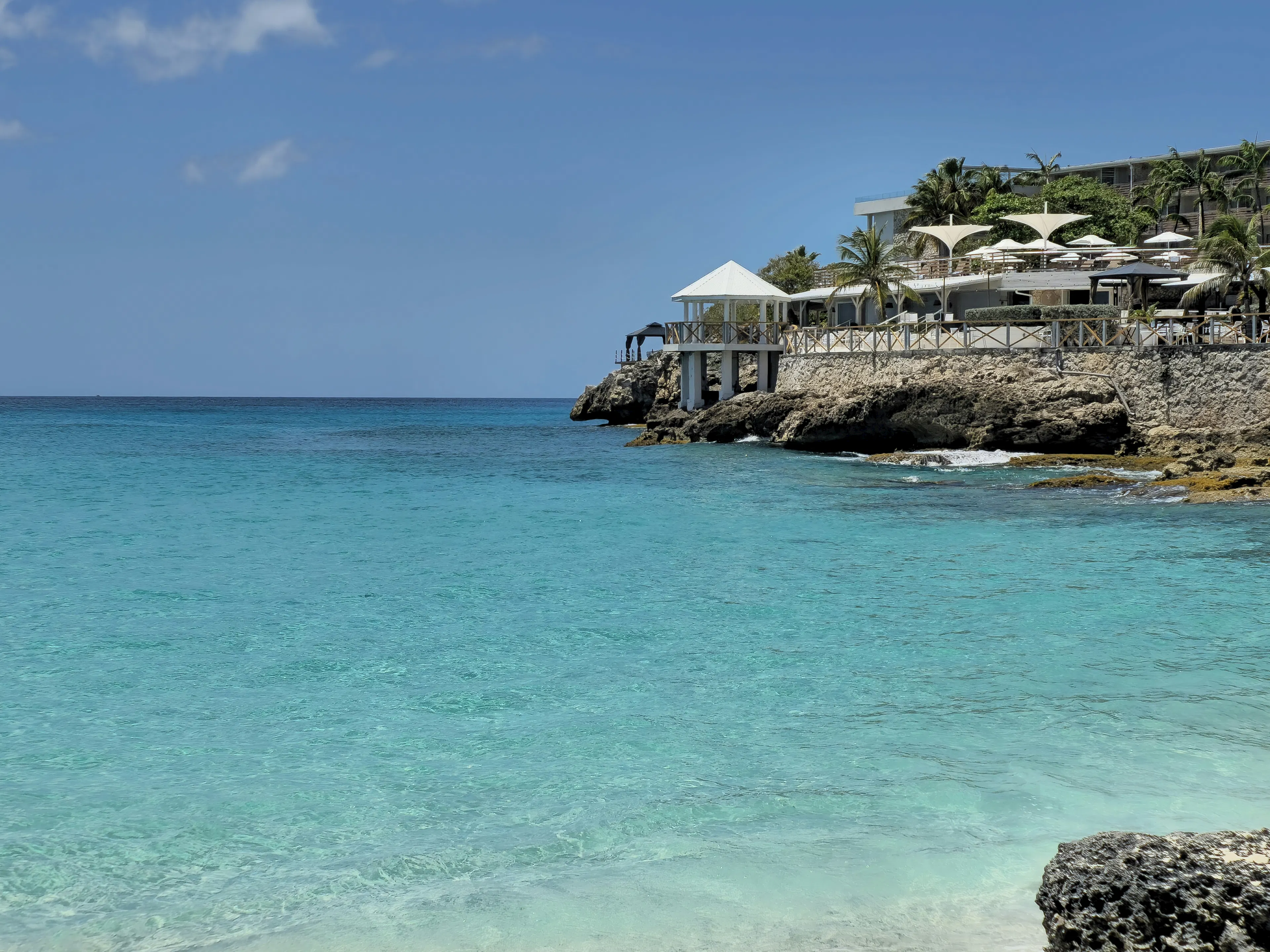 Clear turquoise water beside a rocky shoreline with a waterfront resort and palm trees overlooking the coast.