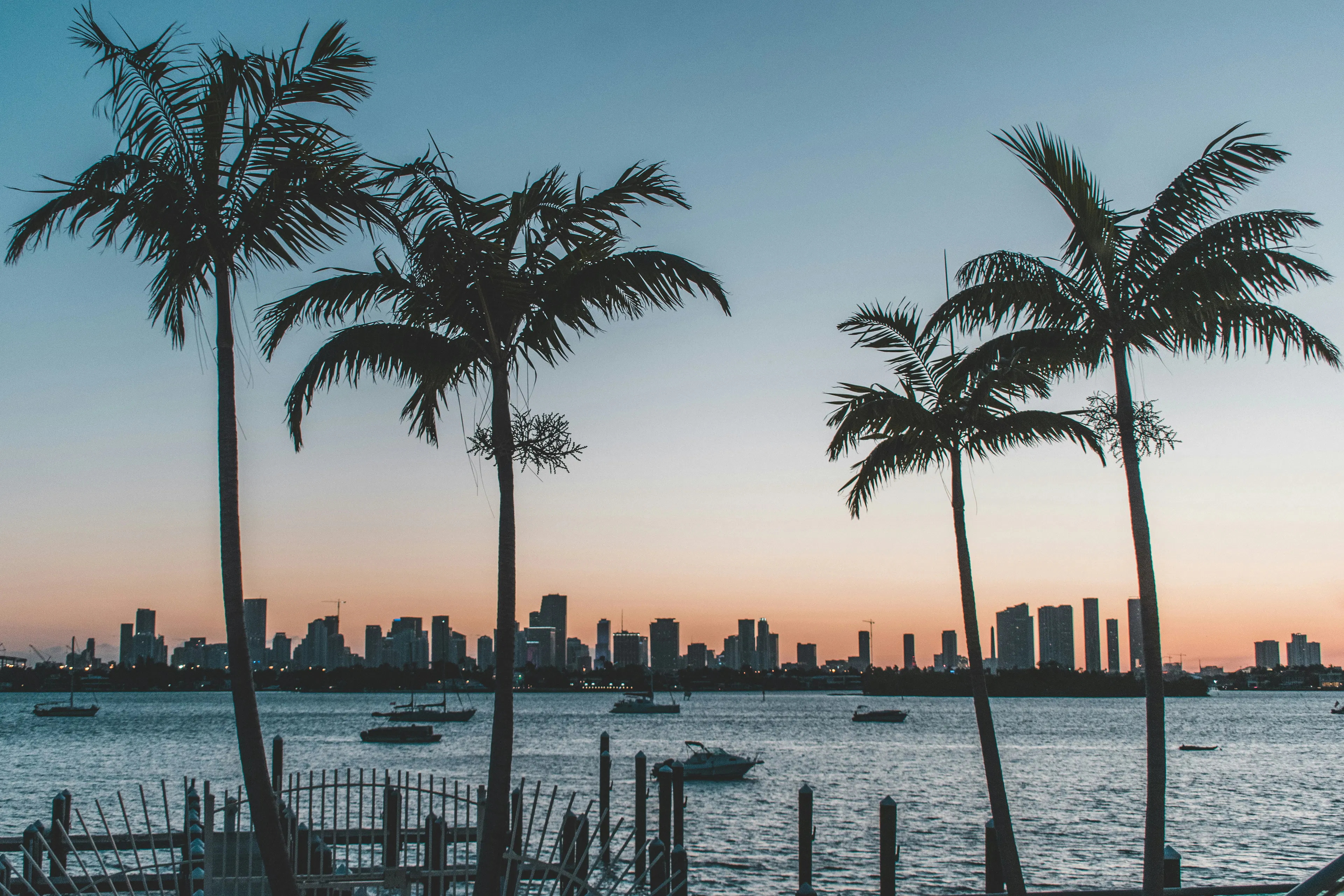 Palm trees and boats along the waterfront with the Miami skyline at sunset in the background.