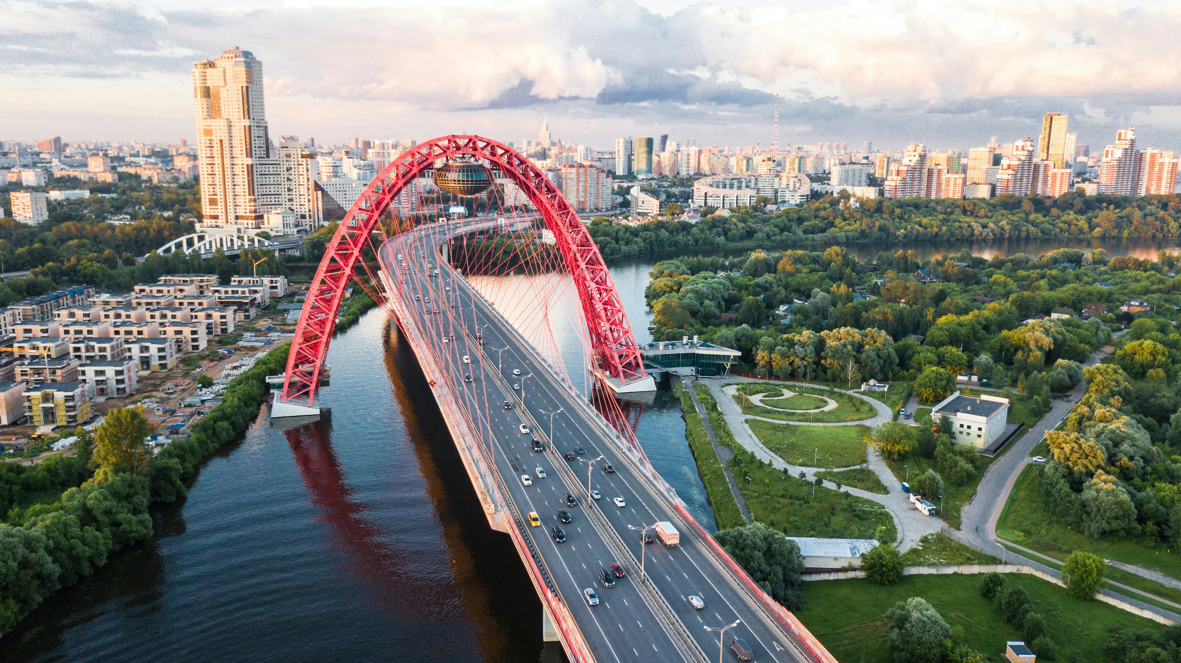 Aerial view of a red arch bridge over a river in Moscow with traffic, trees, and city buildings in the background.