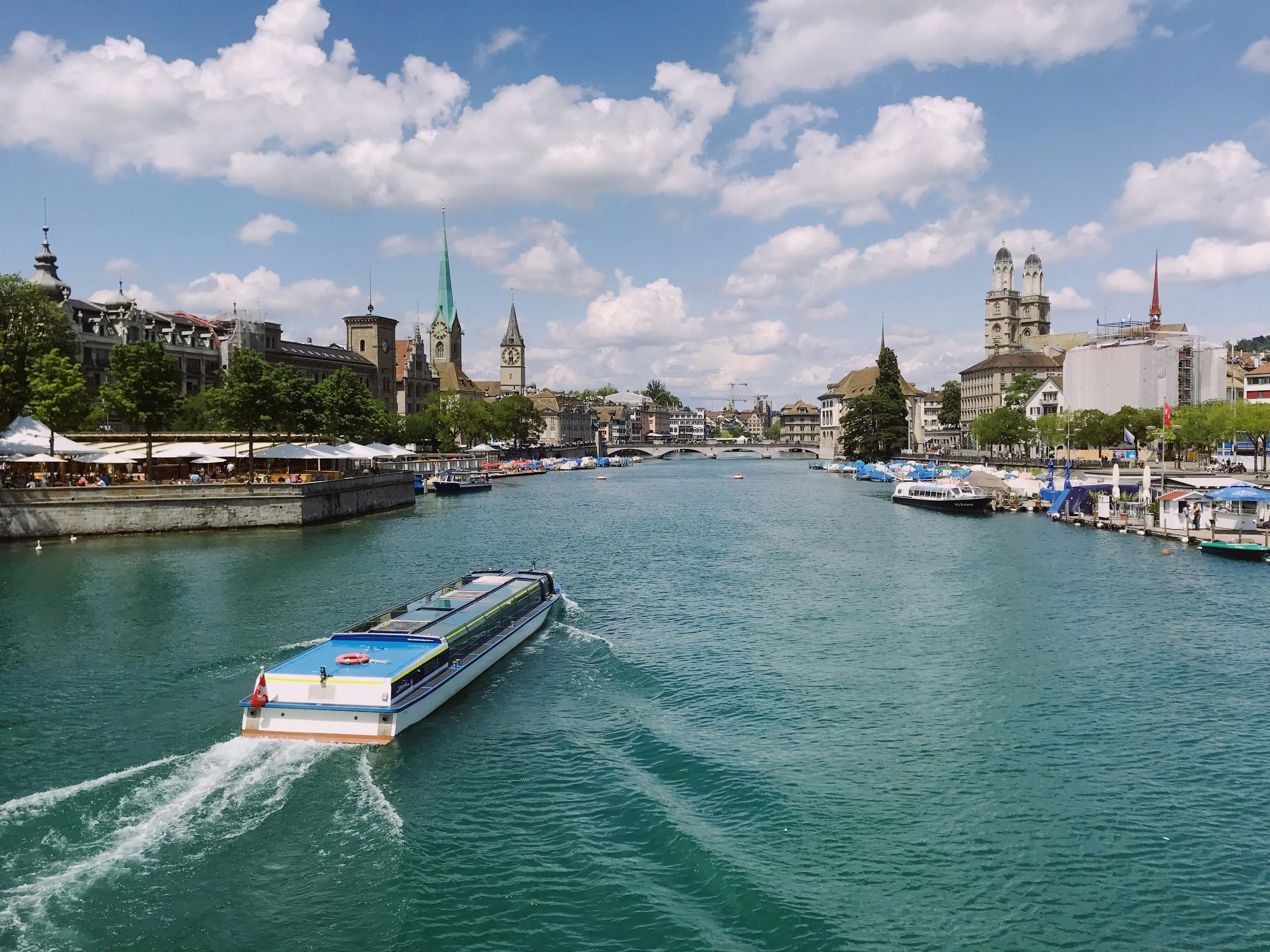 Passenger boat cruising along the Limmat River in Zurich, Switzerland, with historic churches, waterfront buildings, and bridges visible in the distance.