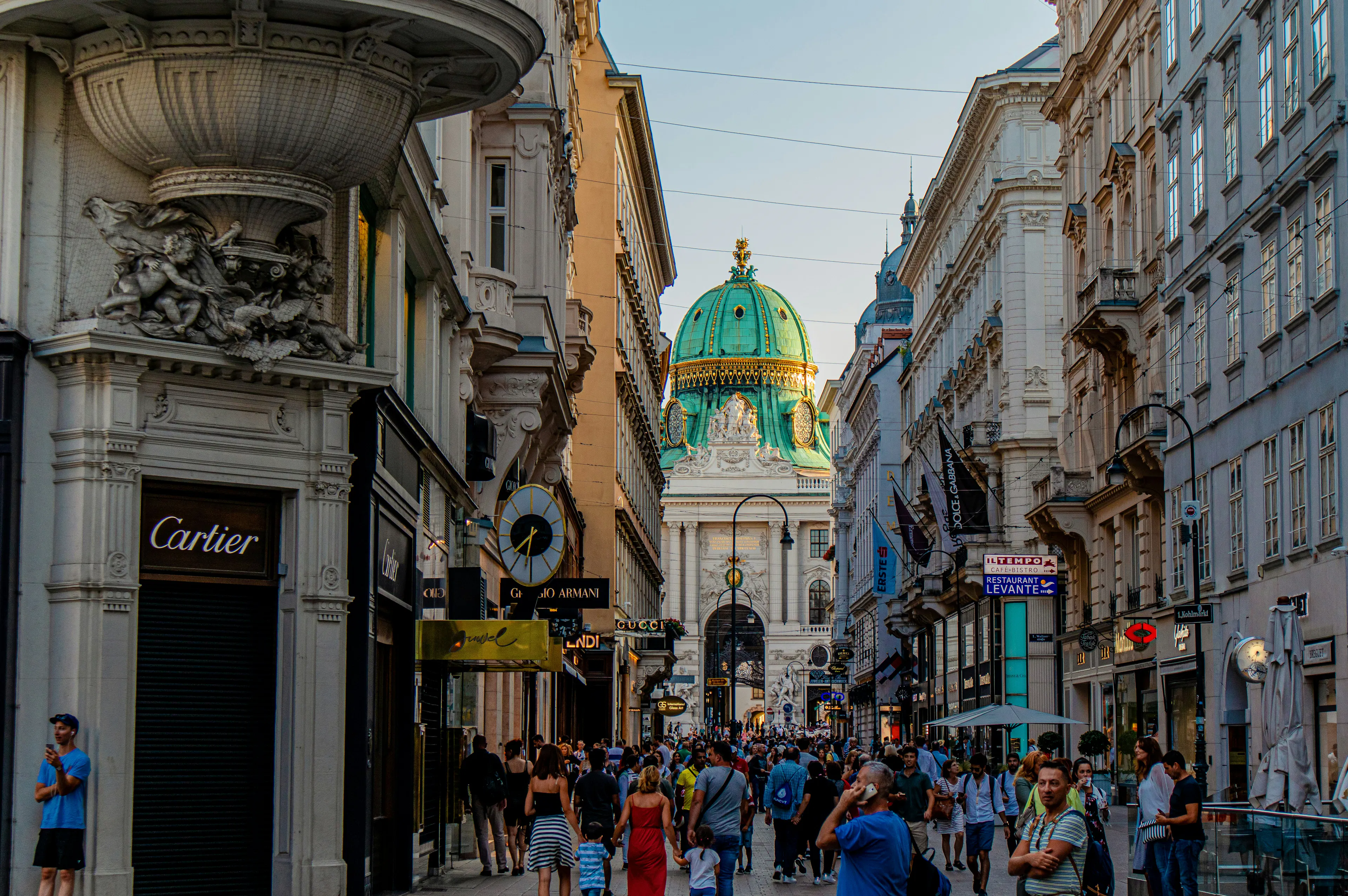 Busy Vienna shopping street with pedestrians, storefronts, historic buildings, and a green-domed building in the distance.
