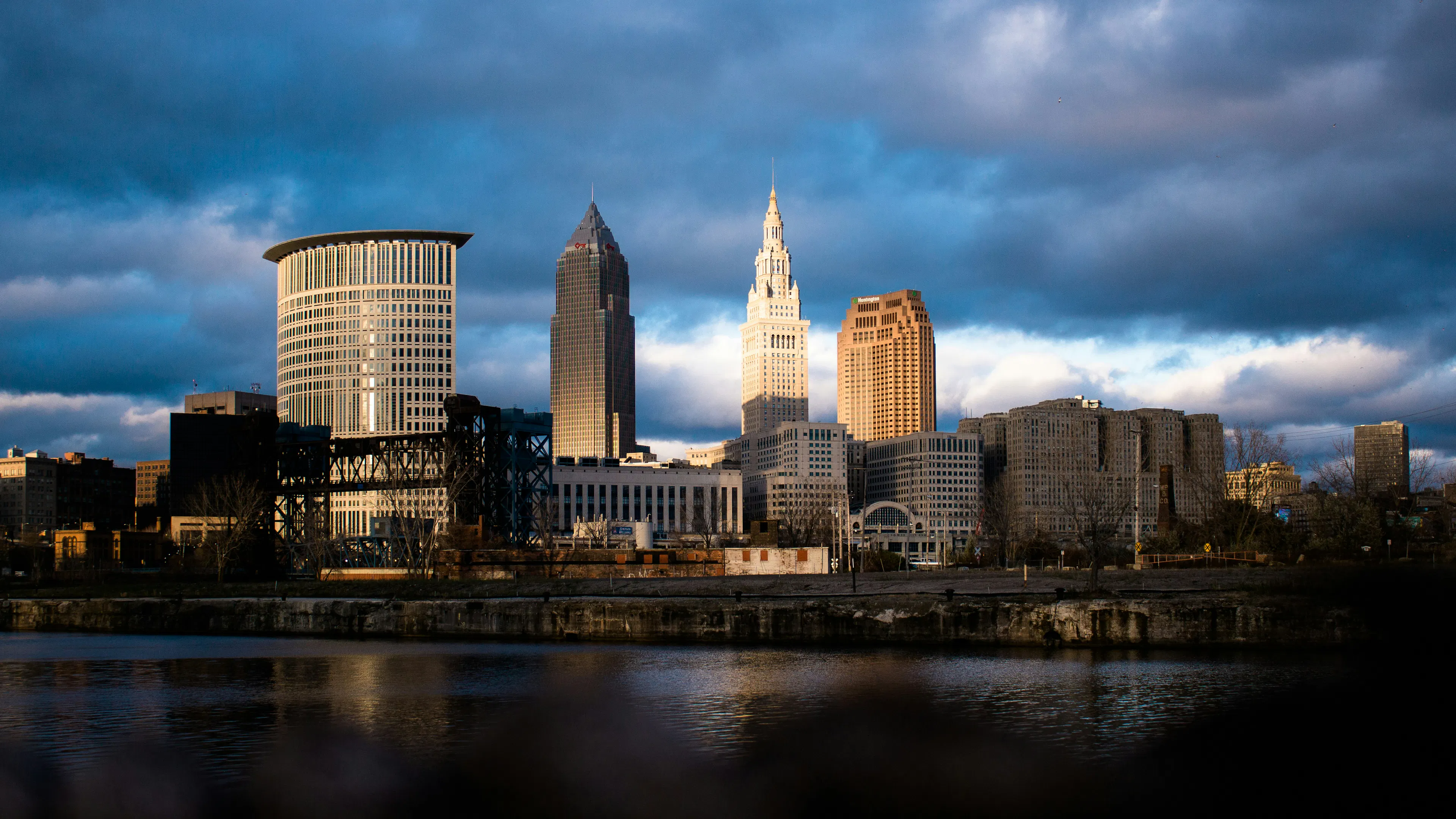 Downtown Cleveland skyline viewed across the river with illuminated skyscrapers beneath dramatic clouds.