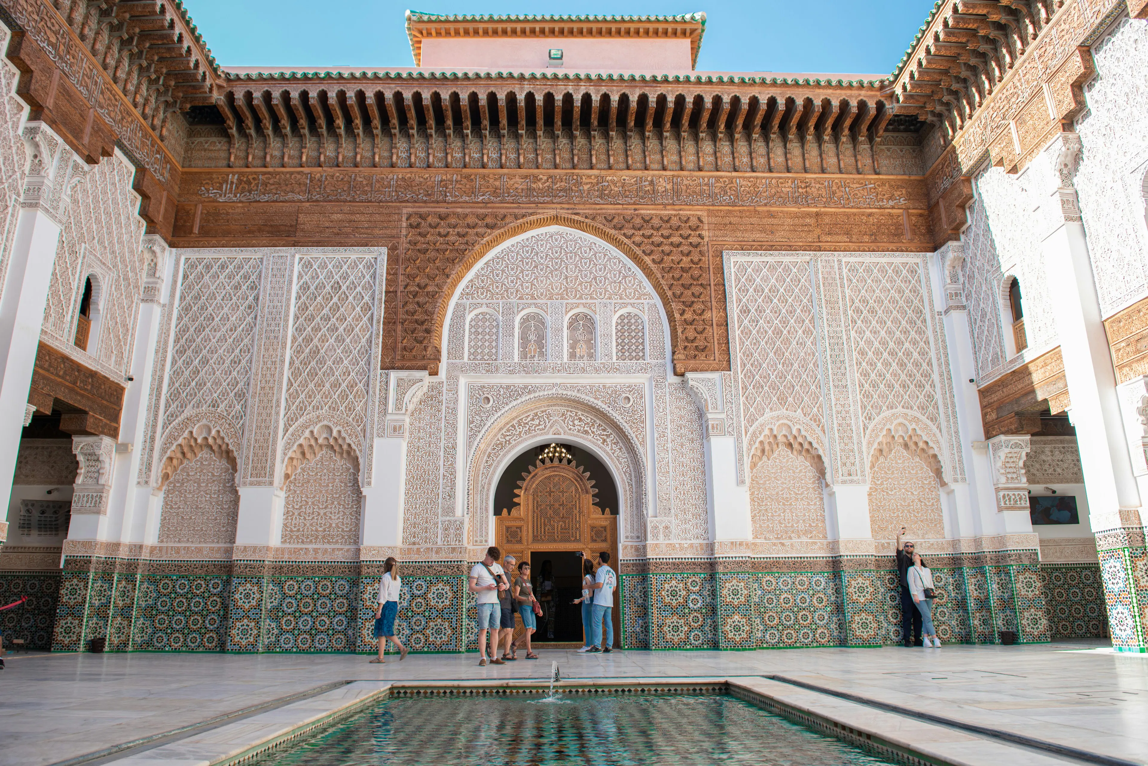Ornately decorated Moroccan courtyard with intricate tilework, carved archways, a reflecting pool, and visitors standing near the entrance.