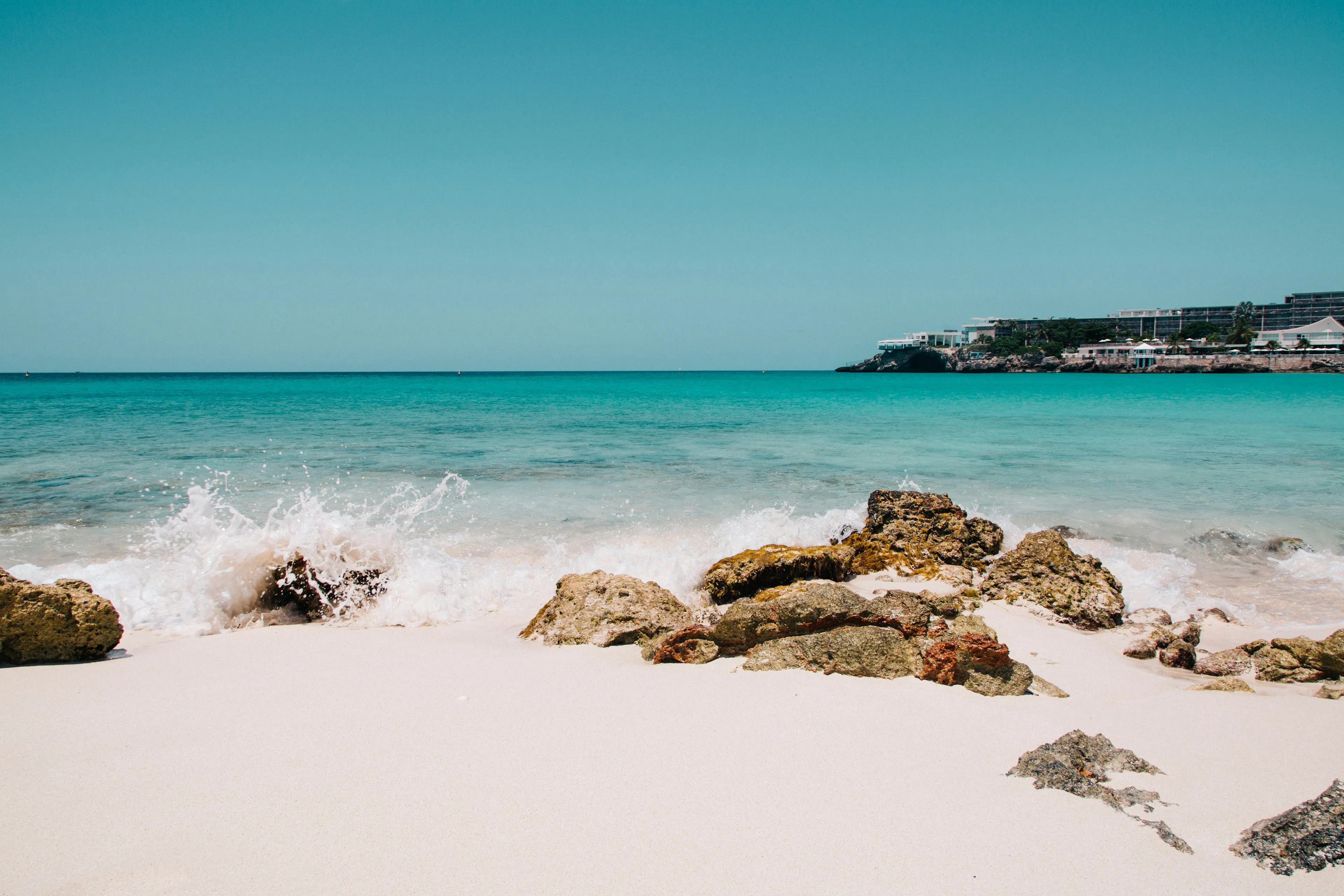 Turquoise ocean waves washing onto a sandy beach with rocky shoreline and coastal buildings in the distance.