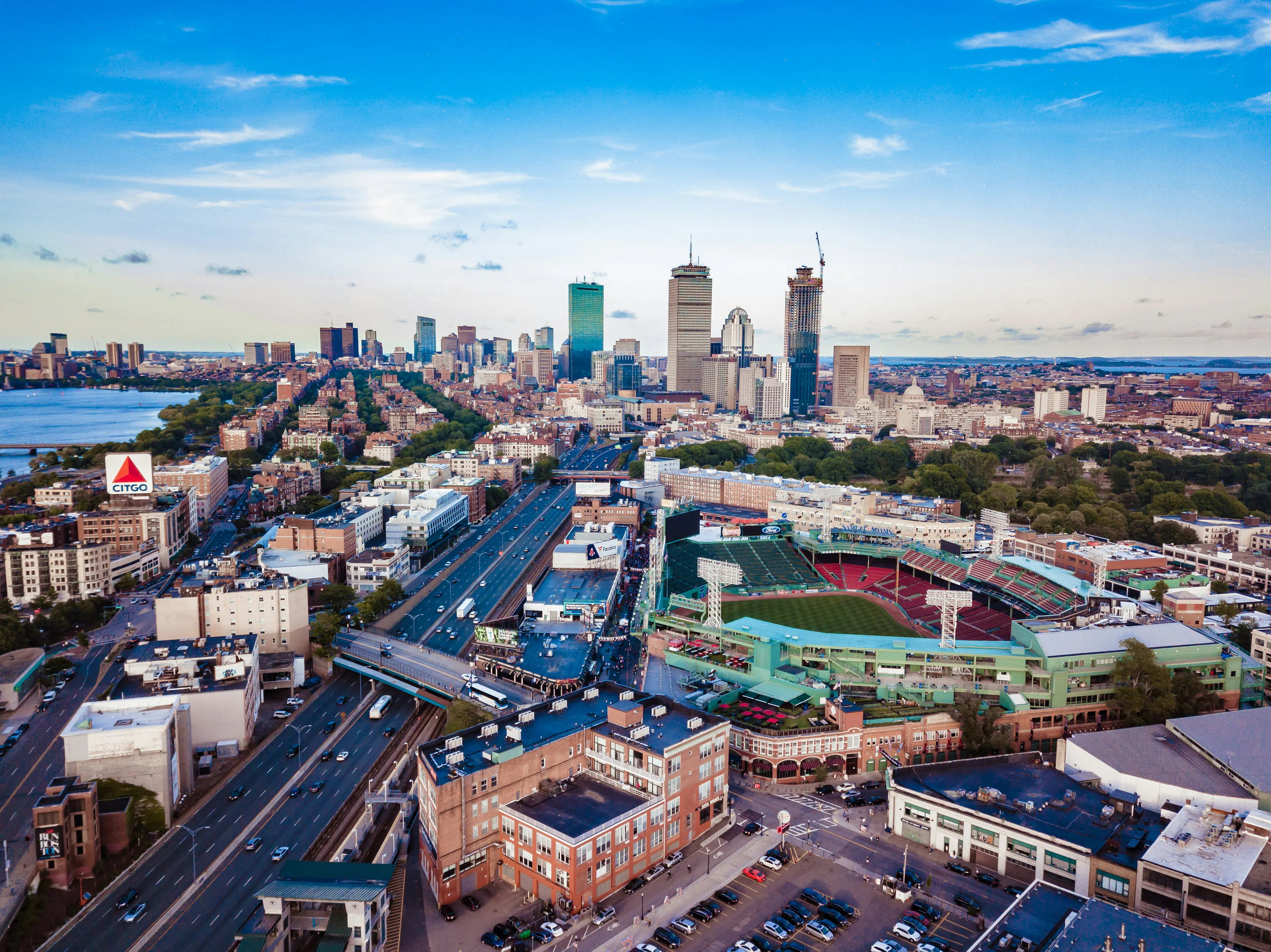 Aerial view of Boston with Fenway Park, downtown skyscrapers, and major roadways extending across the city.