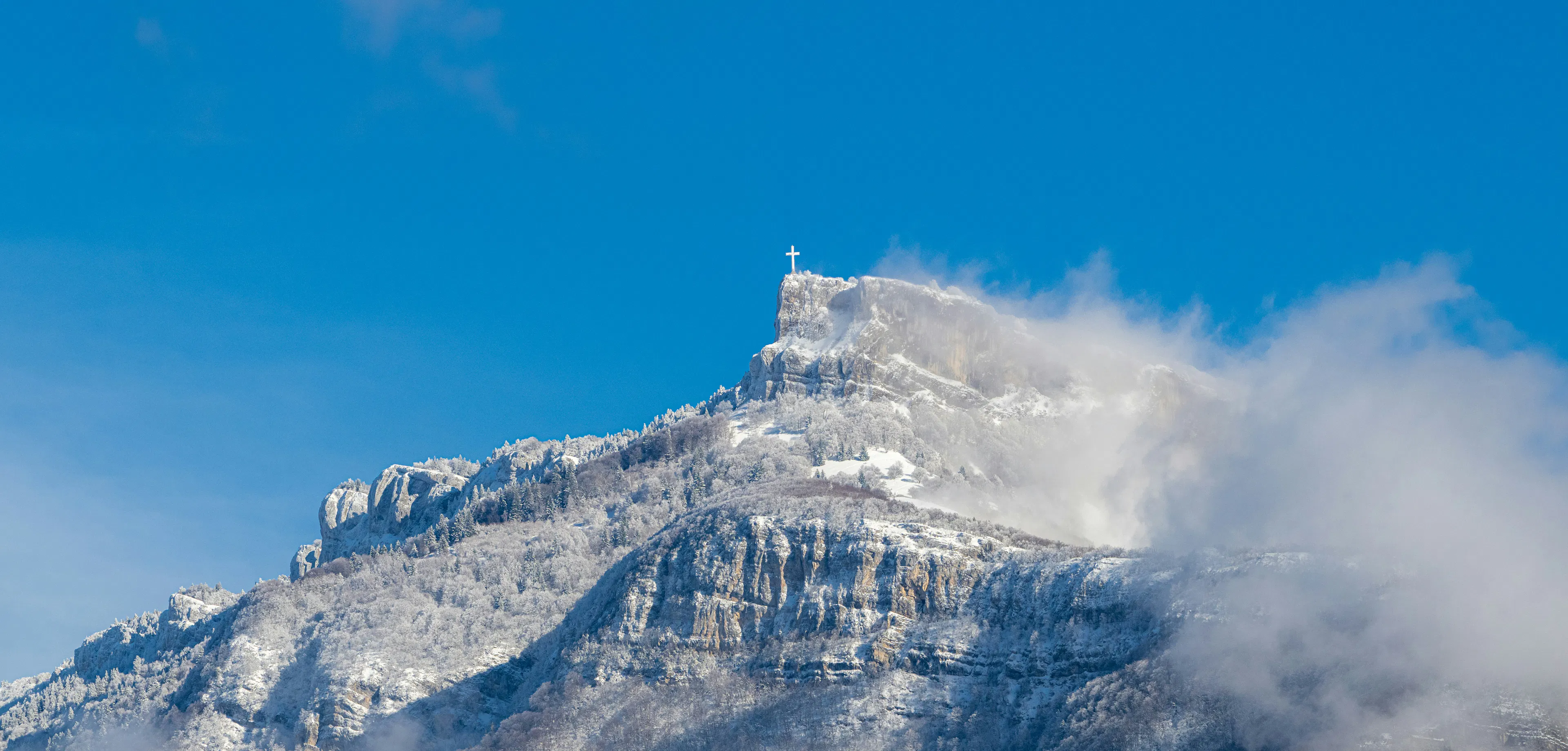 Snow-covered mountain peak with a cross at the summit under a clear blue sky and passing clouds.