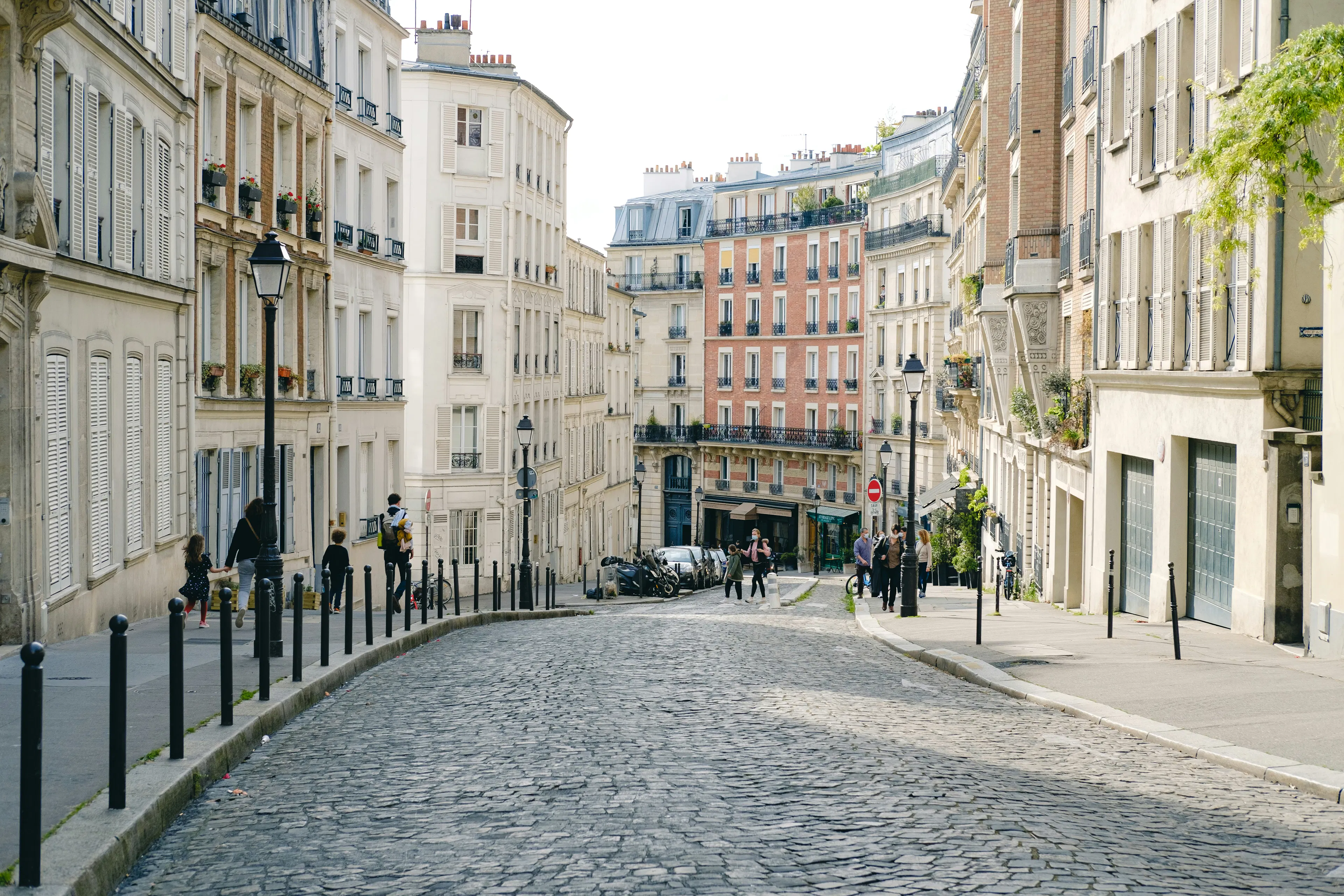 Cobblestone street in Paris lined with historic apartment buildings, parked vehicles, and pedestrians.