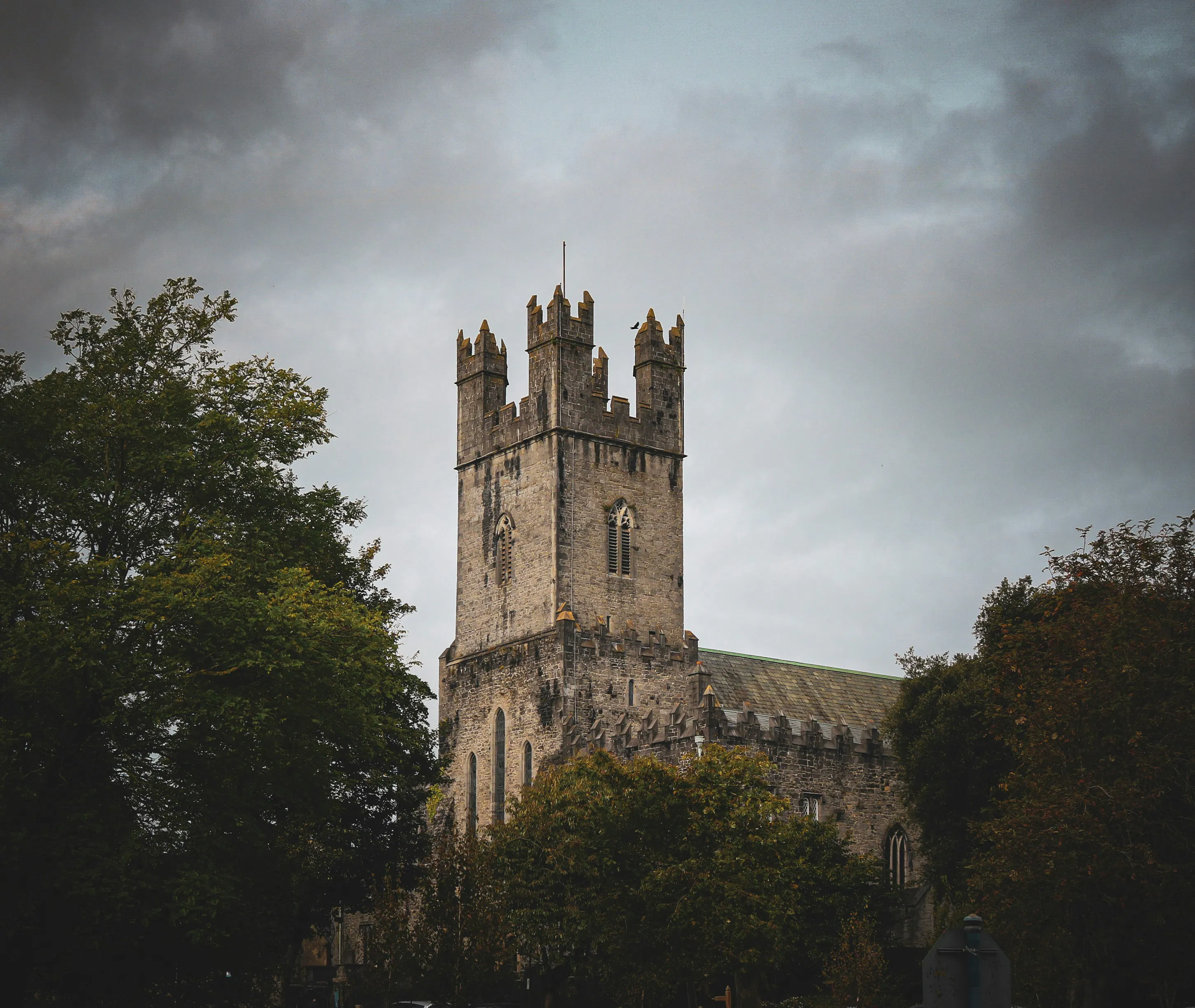 Historic stone church with a square tower rising above trees under an overcast sky.