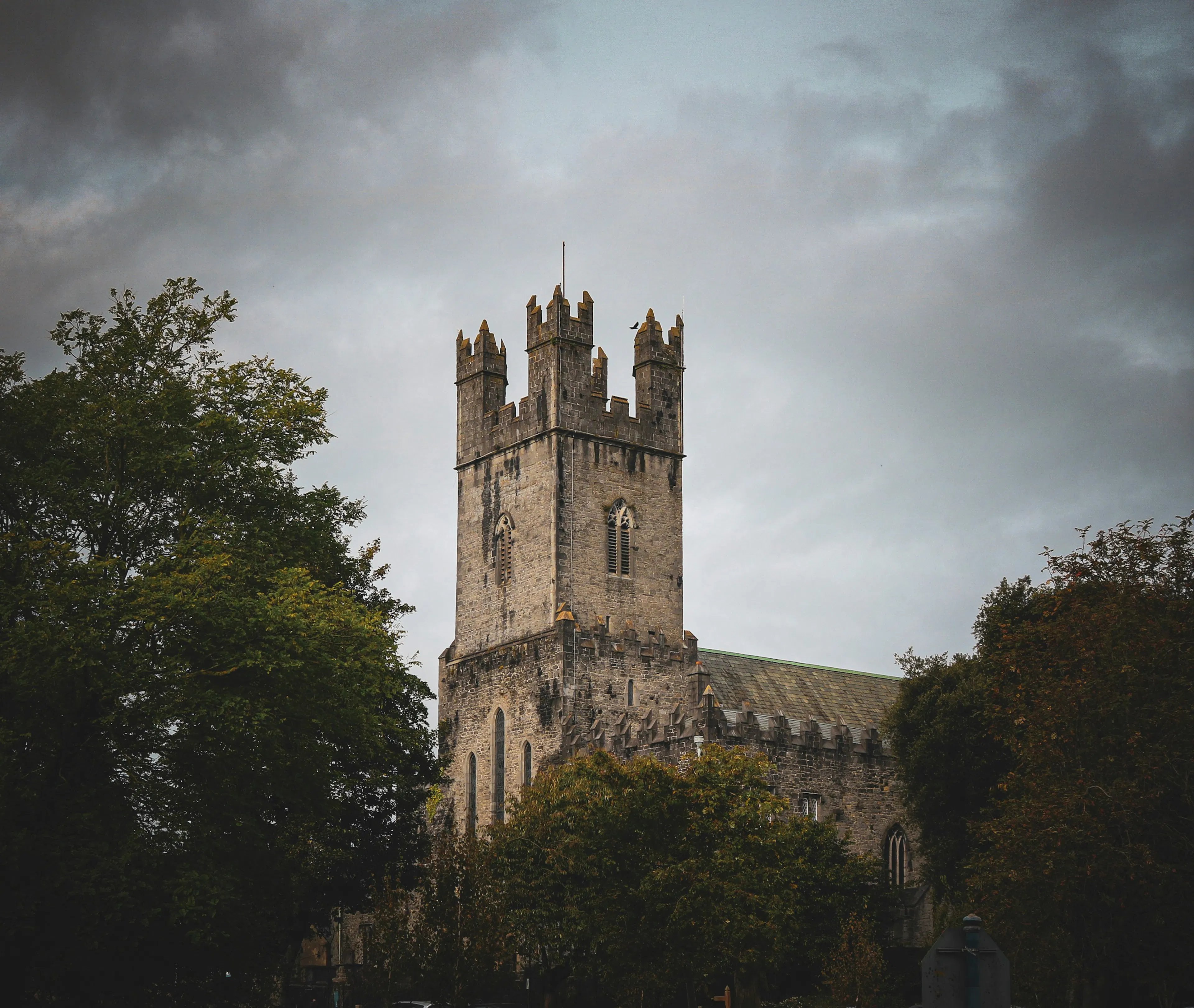 Historic stone church with a square tower rising above trees under an overcast sky.