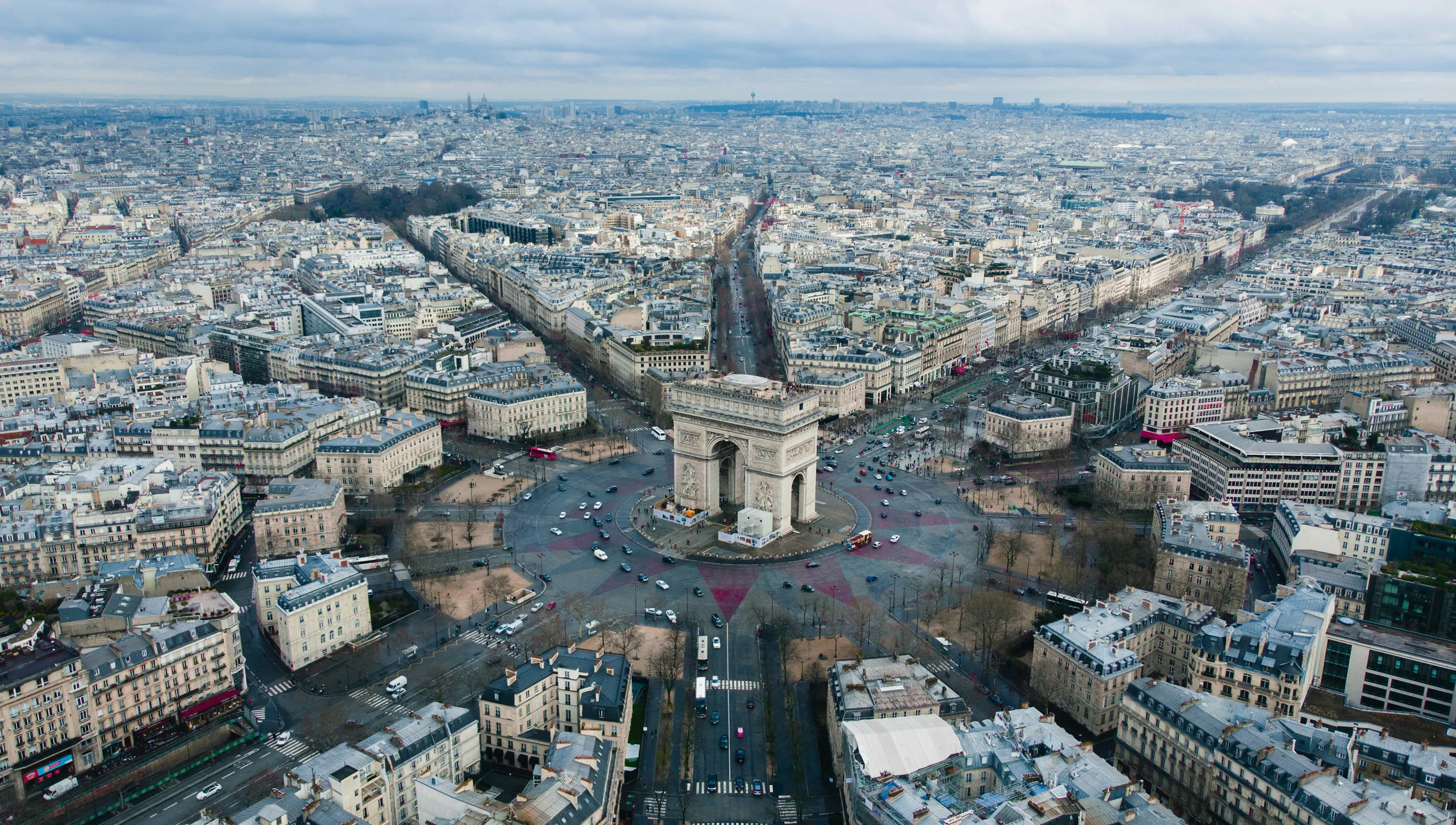 Aerial view of the Arc de Triomphe at the center of Paris with radiating avenues and surrounding city blocks.