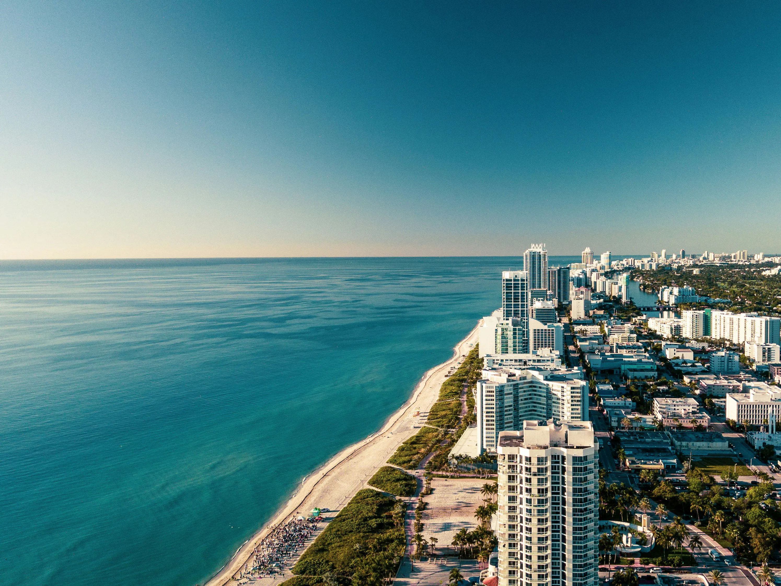 Aerial view of Miami Beach with oceanfront high-rise buildings, sandy shoreline, and turquoise water stretching along the coast.