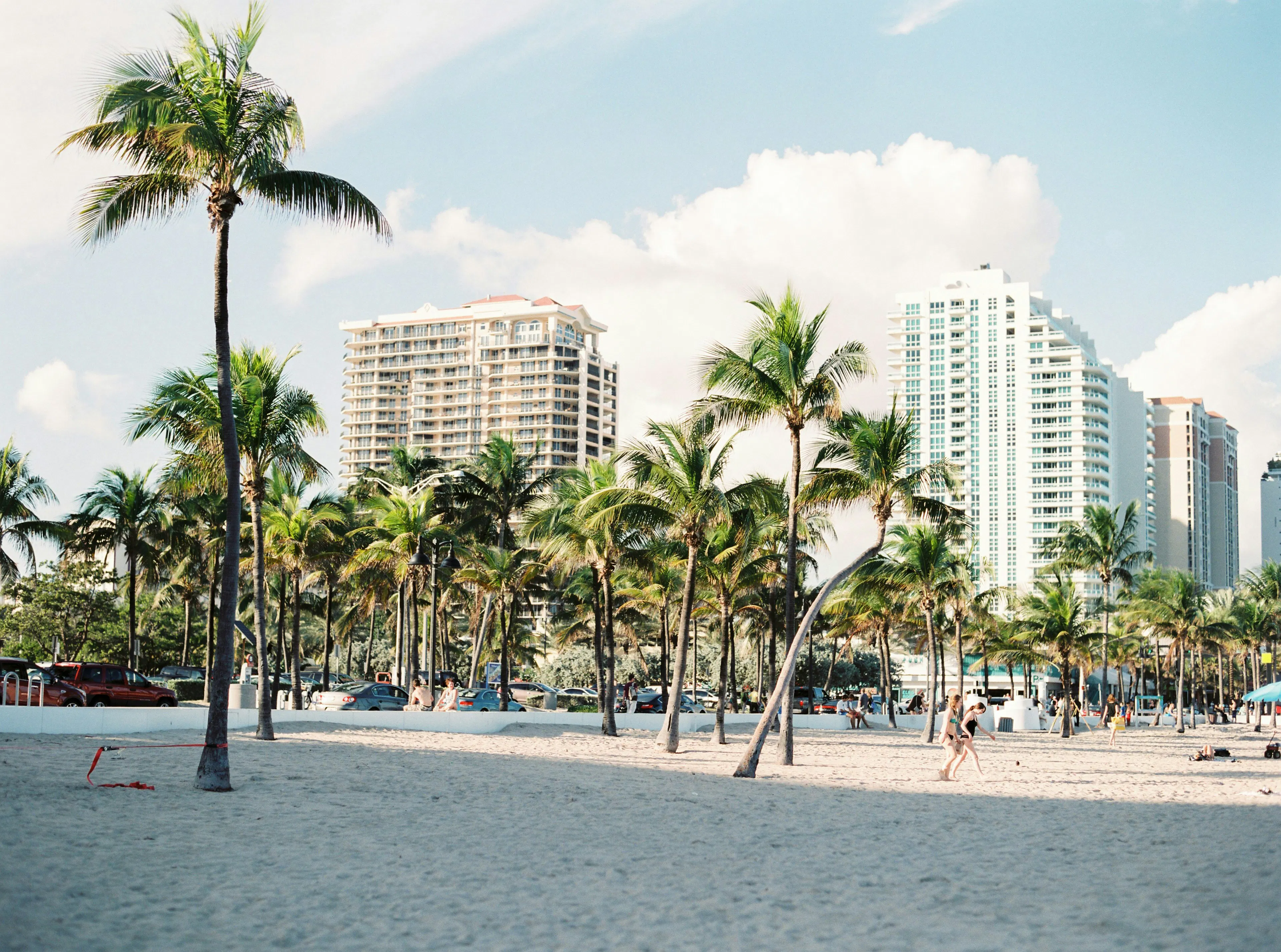Sandy beach lined with palm trees, with high-rise buildings and a few people visible in the background under a sunny sky.