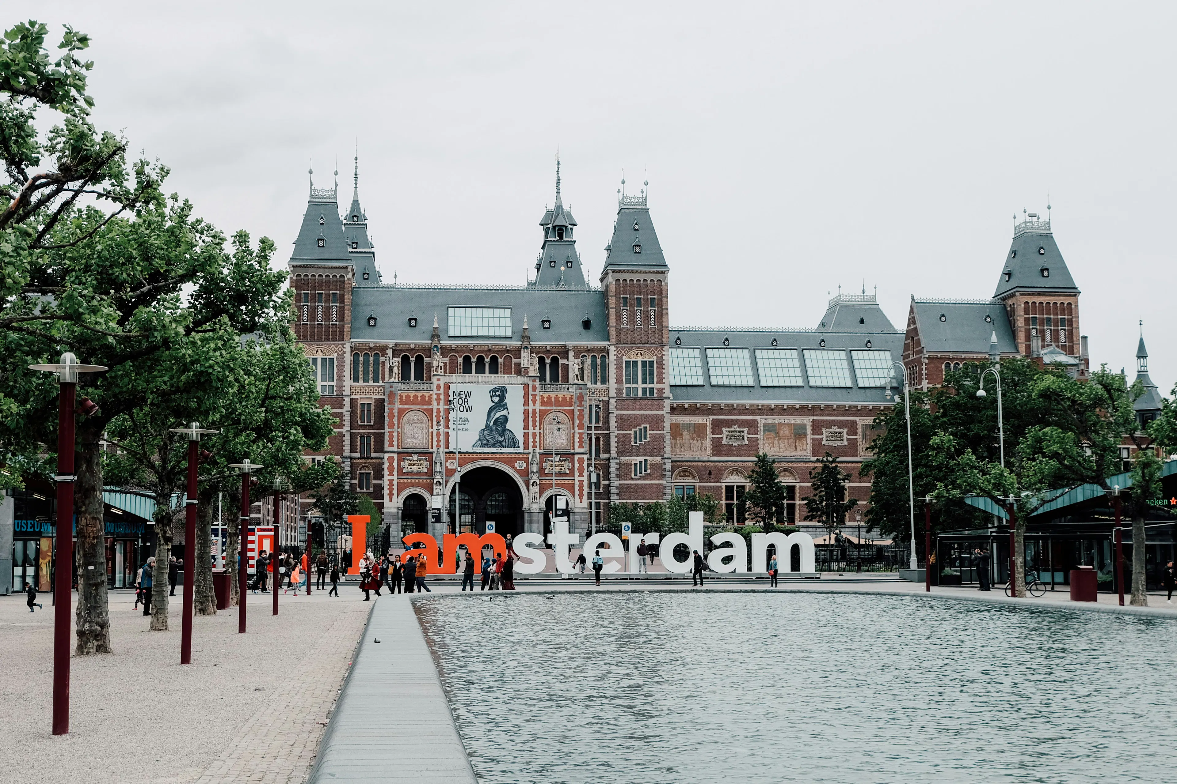 View of the Rijksmuseum and the iconic “I amsterdam” sign in a public square, framed by trees and historic architecture.