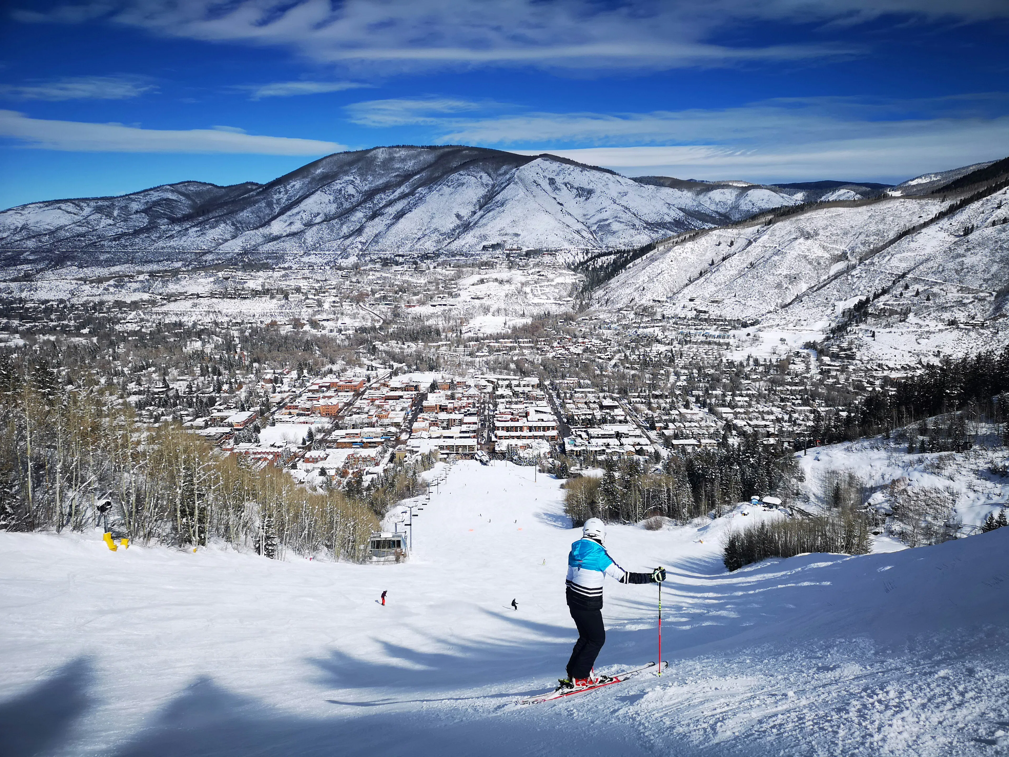 Skier descending a snow-covered slope overlooking Aspen and surrounding mountain peaks under a clear blue sky.
