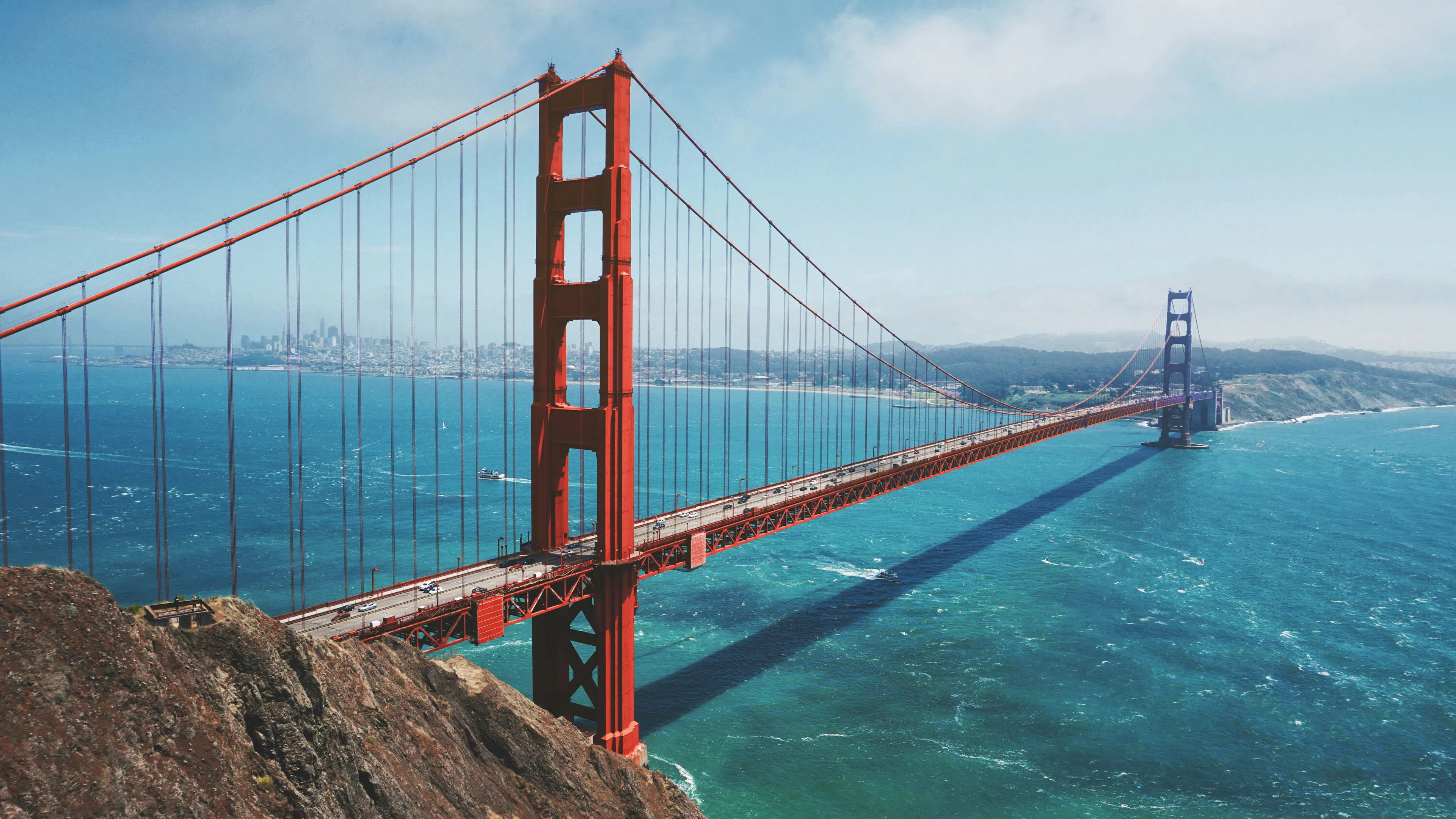 Golden Gate Bridge spanning San Francisco Bay with blue water, coastal cliffs, and the city skyline in the distance.