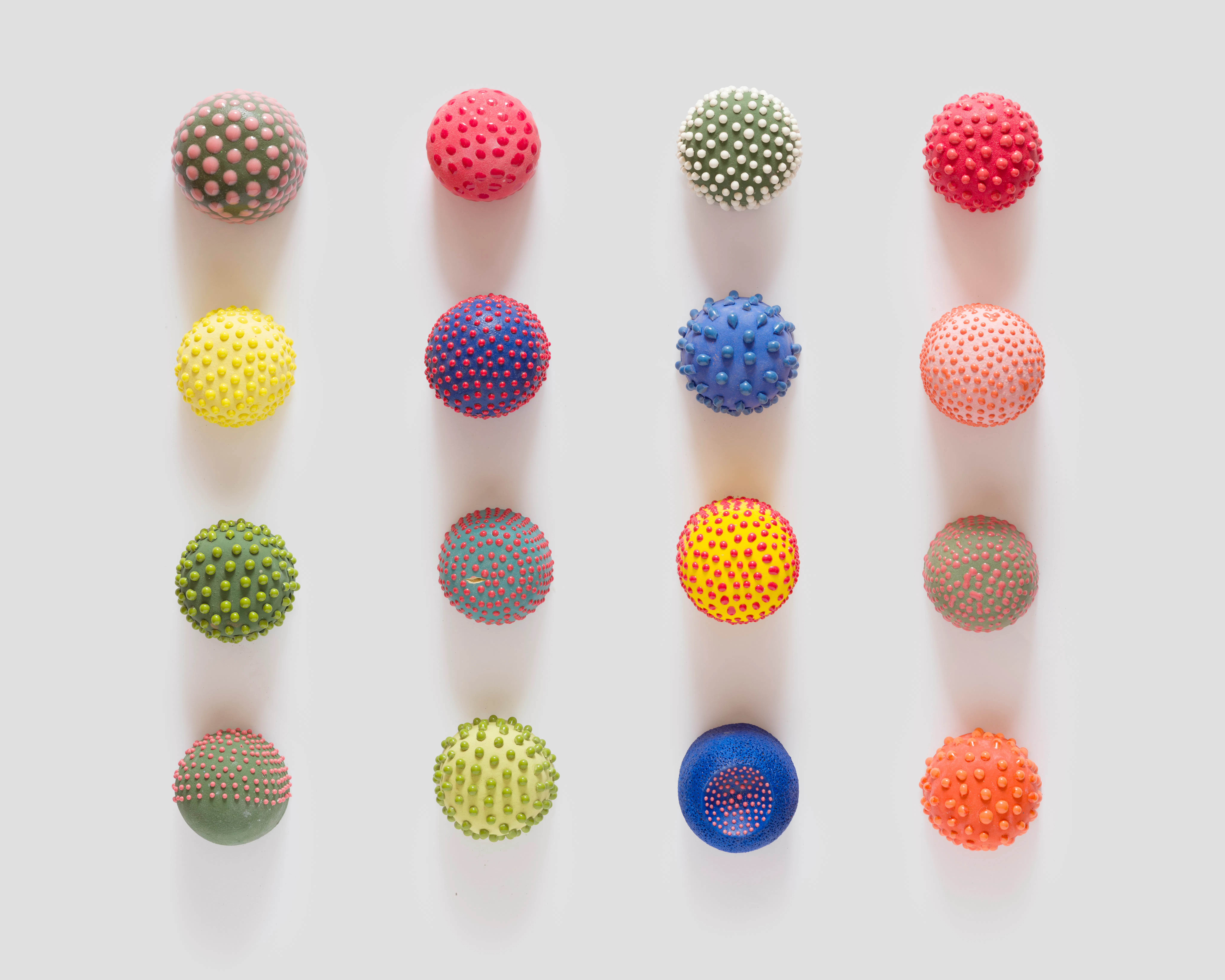 Sixteen small ceramic seed forms by Theo Hirschfield arranged in four rows, featuring varied bright colors, glazes, and raised surface textures.