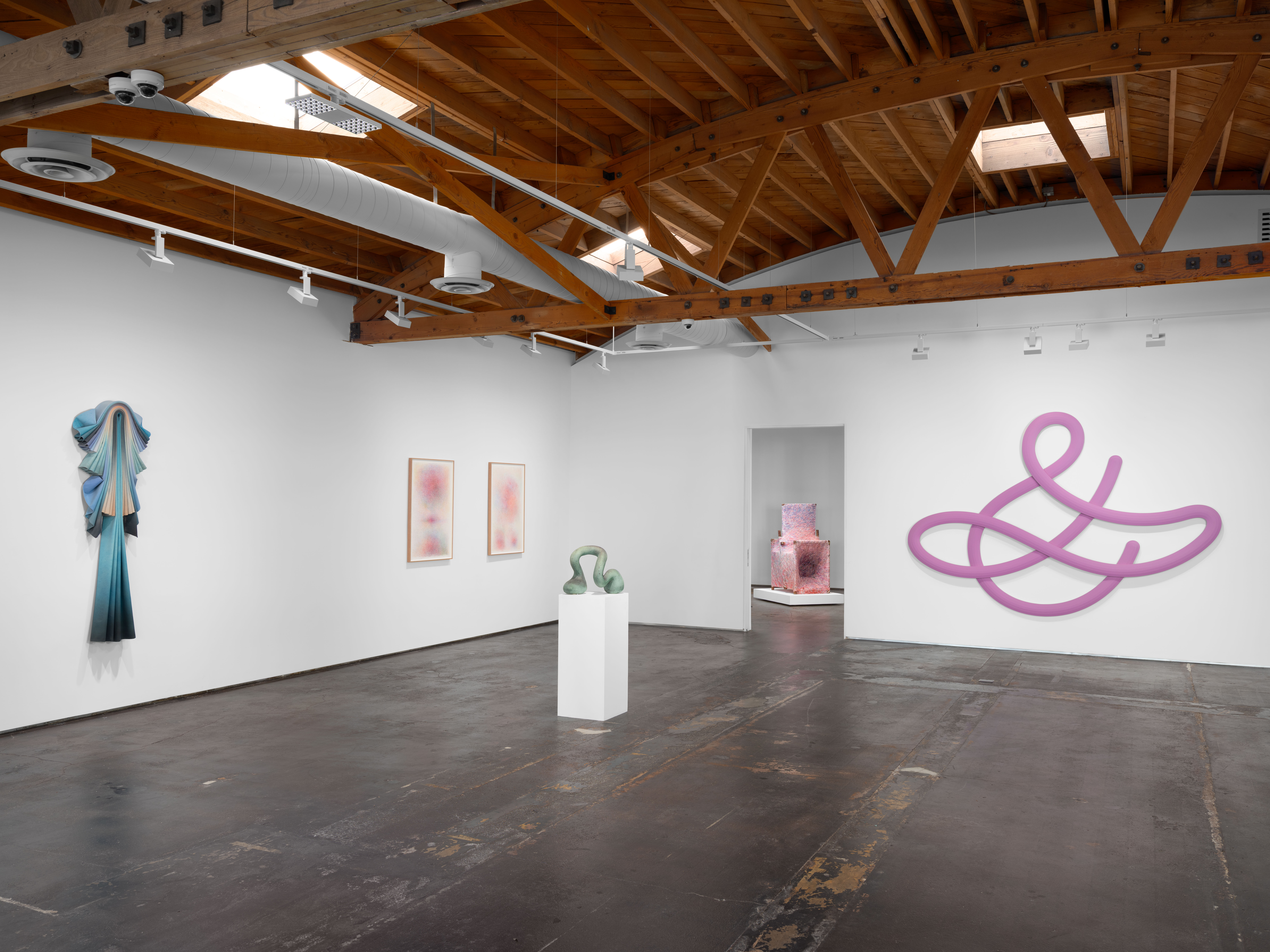 A wide view of a gallery interior shows multiple artworks installed on white walls beneath exposed wooden ceiling beams.