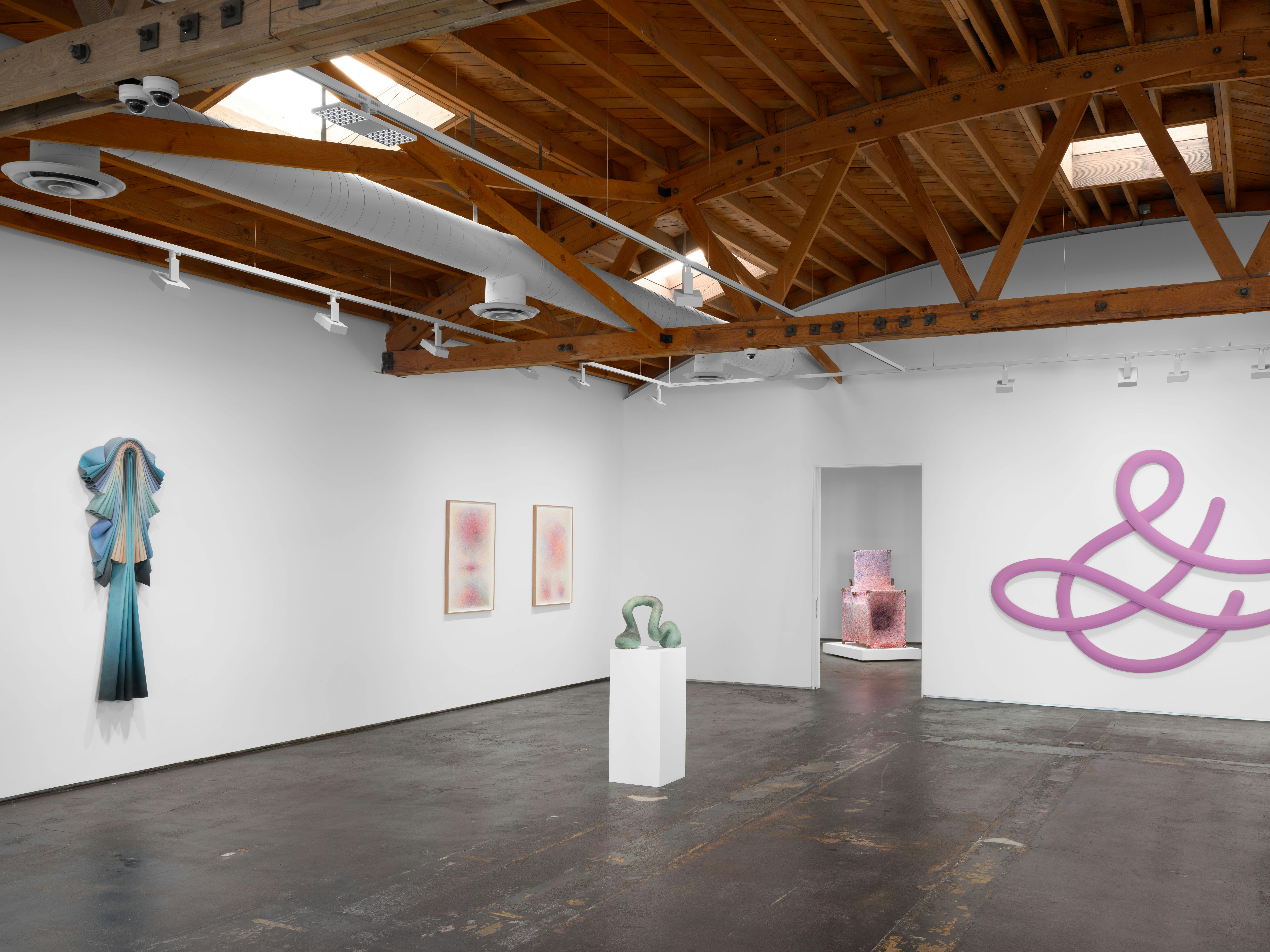 A wide view of a gallery interior shows multiple artworks installed on white walls beneath exposed wooden ceiling beams.