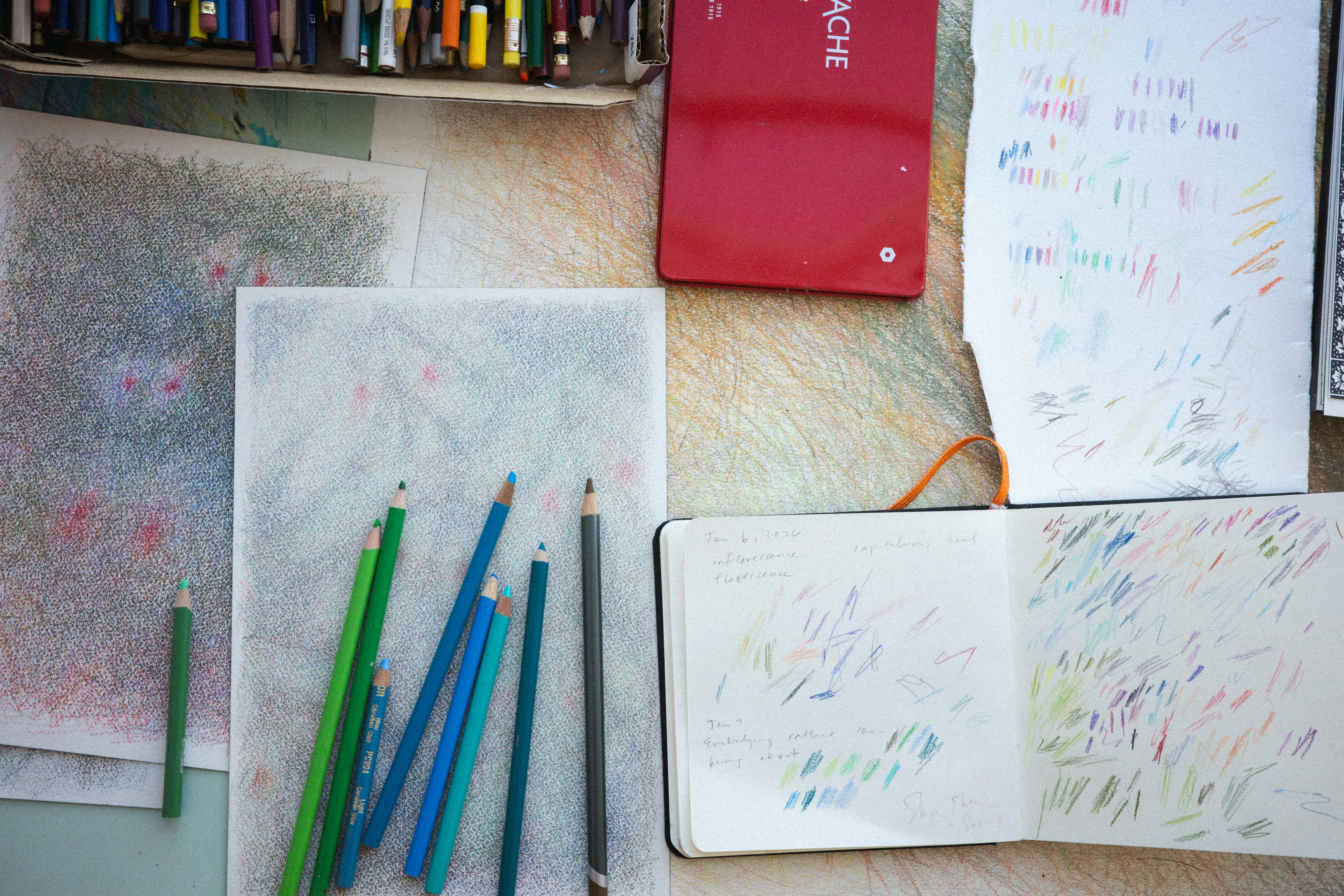 Overhead view of colored pencils and sketchbooks with abstract color studies laid out on a studio work surface.