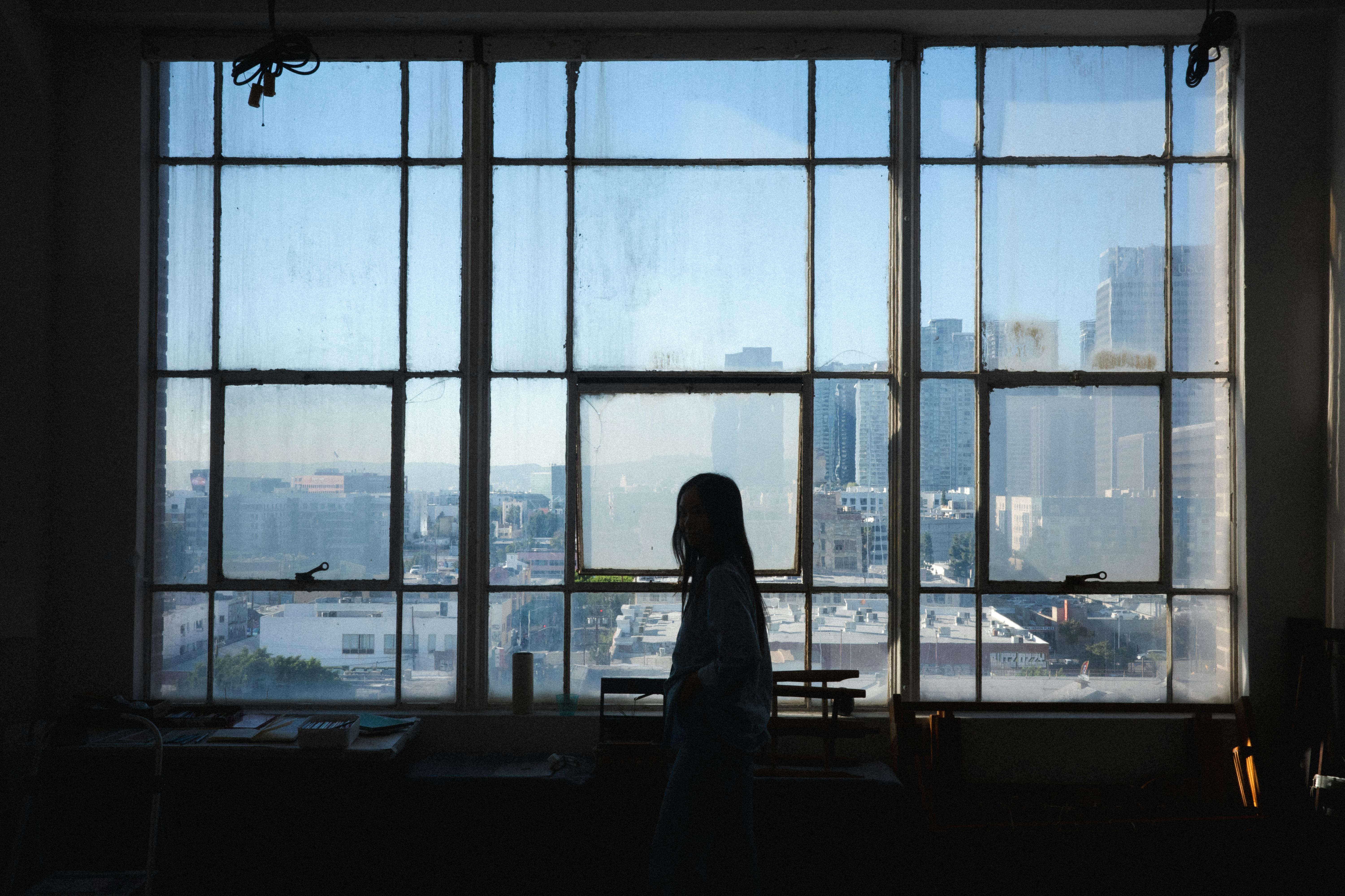 Silhouette of Sheng Lor standing inside her studio, looking out through large windows toward the Los Angeles skyline.