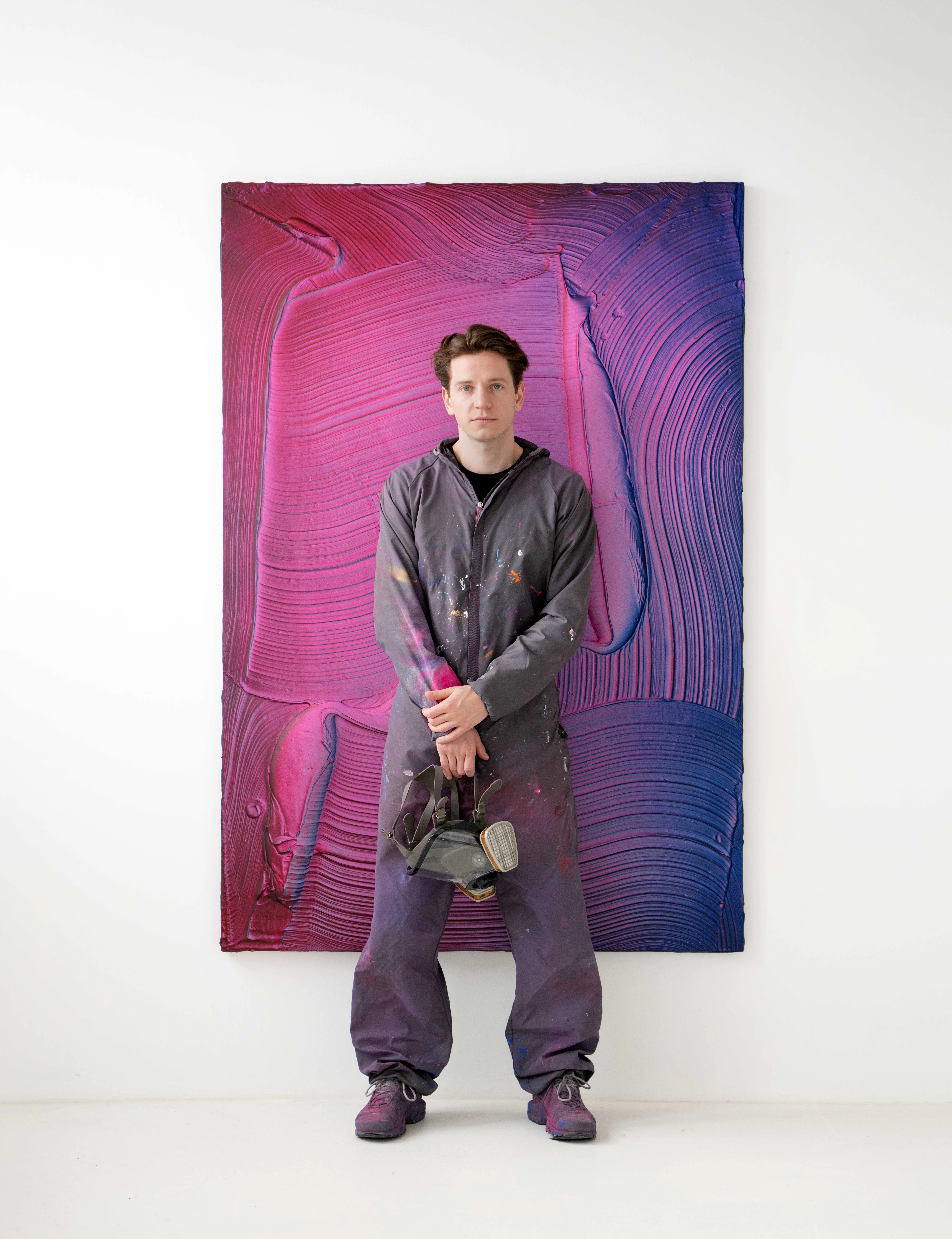 Ruben Benjamin standing in his studio before a textured abstract painting, wearing paint-marked workwear