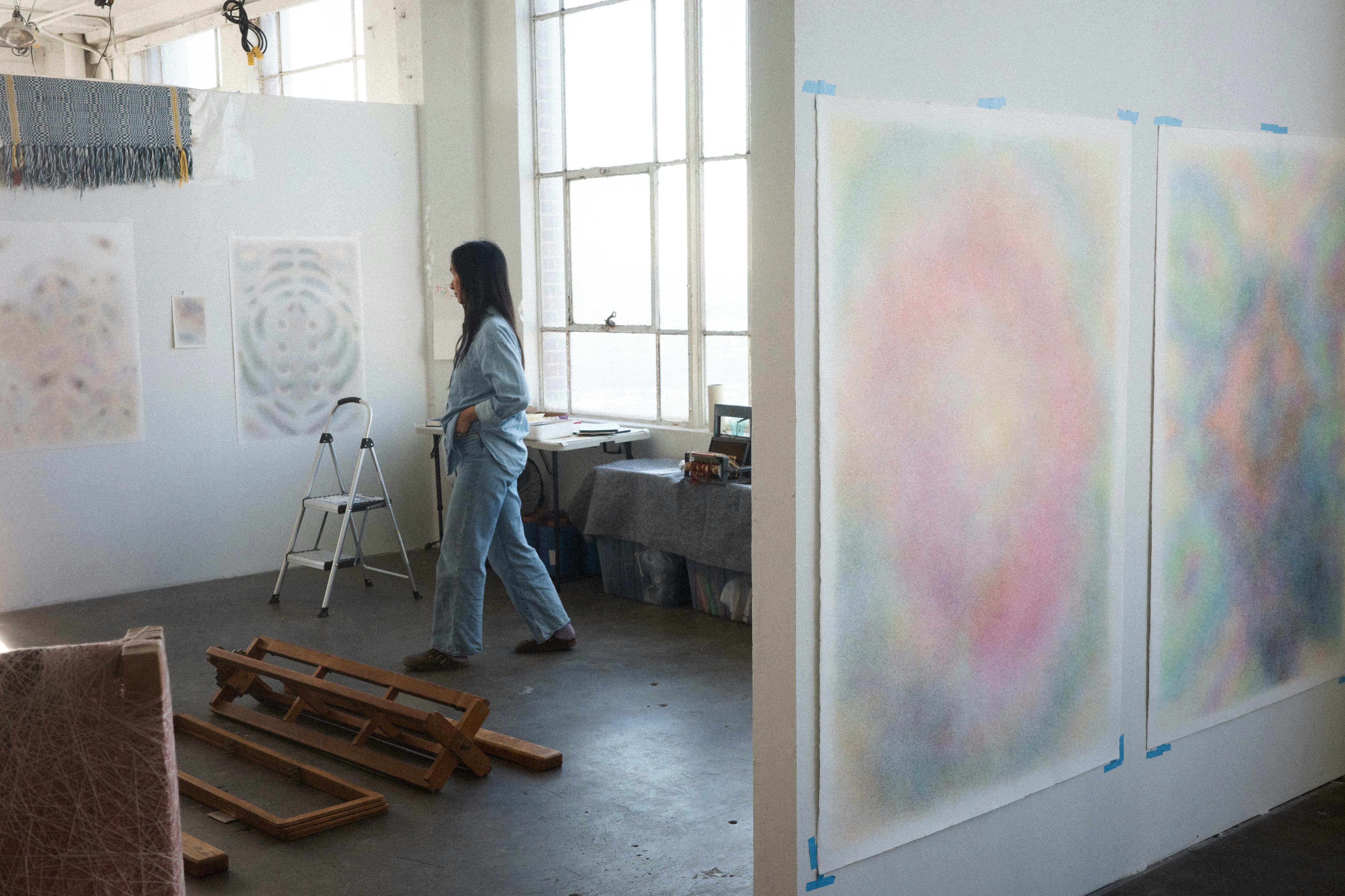 Sheng Lor walking through her studio with large pastel thread drawings mounted on the walls and loom parts on the floor.