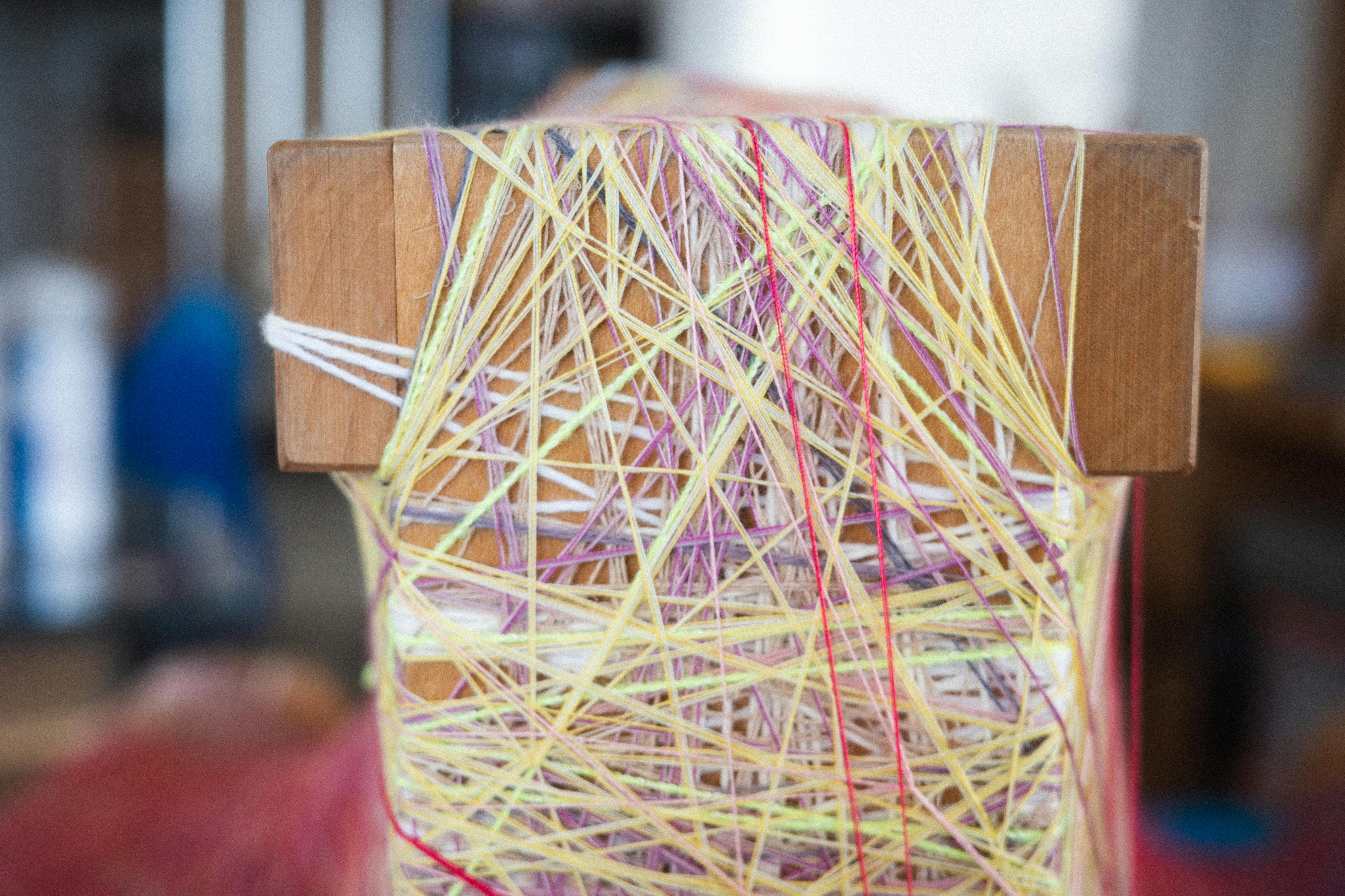 Detail of a wooden loom by Sheng Lor, wrapped with layered pastel threads in yellow, pink, lavender, and white.
