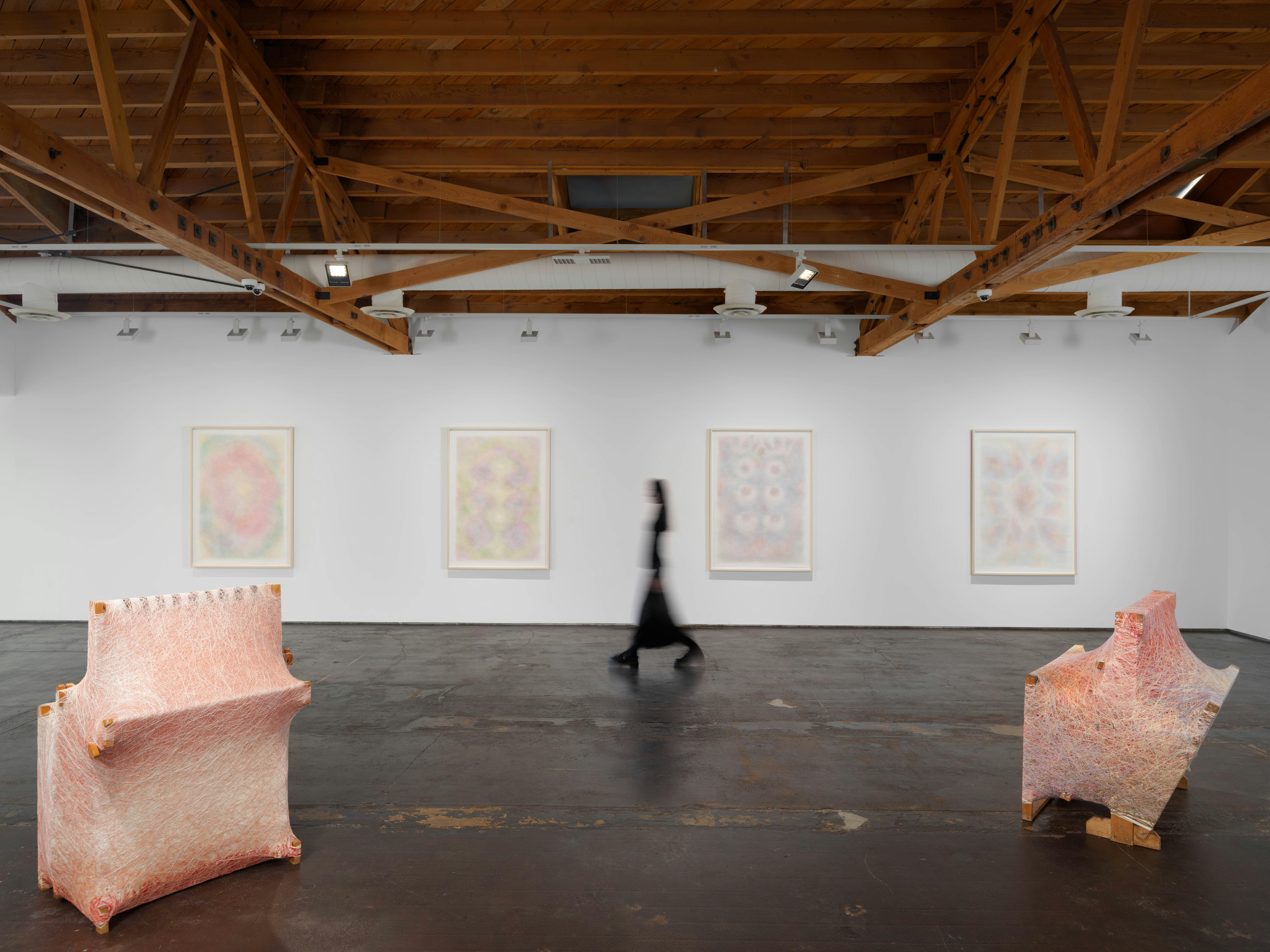 Wide installation view of Interstitial, a solo exhibition by Sheng Lor at Gallery Sonder, featuring pastel-toned wall works and sculptural forms in a white gallery space, with a blurred figure walking through for scale.
