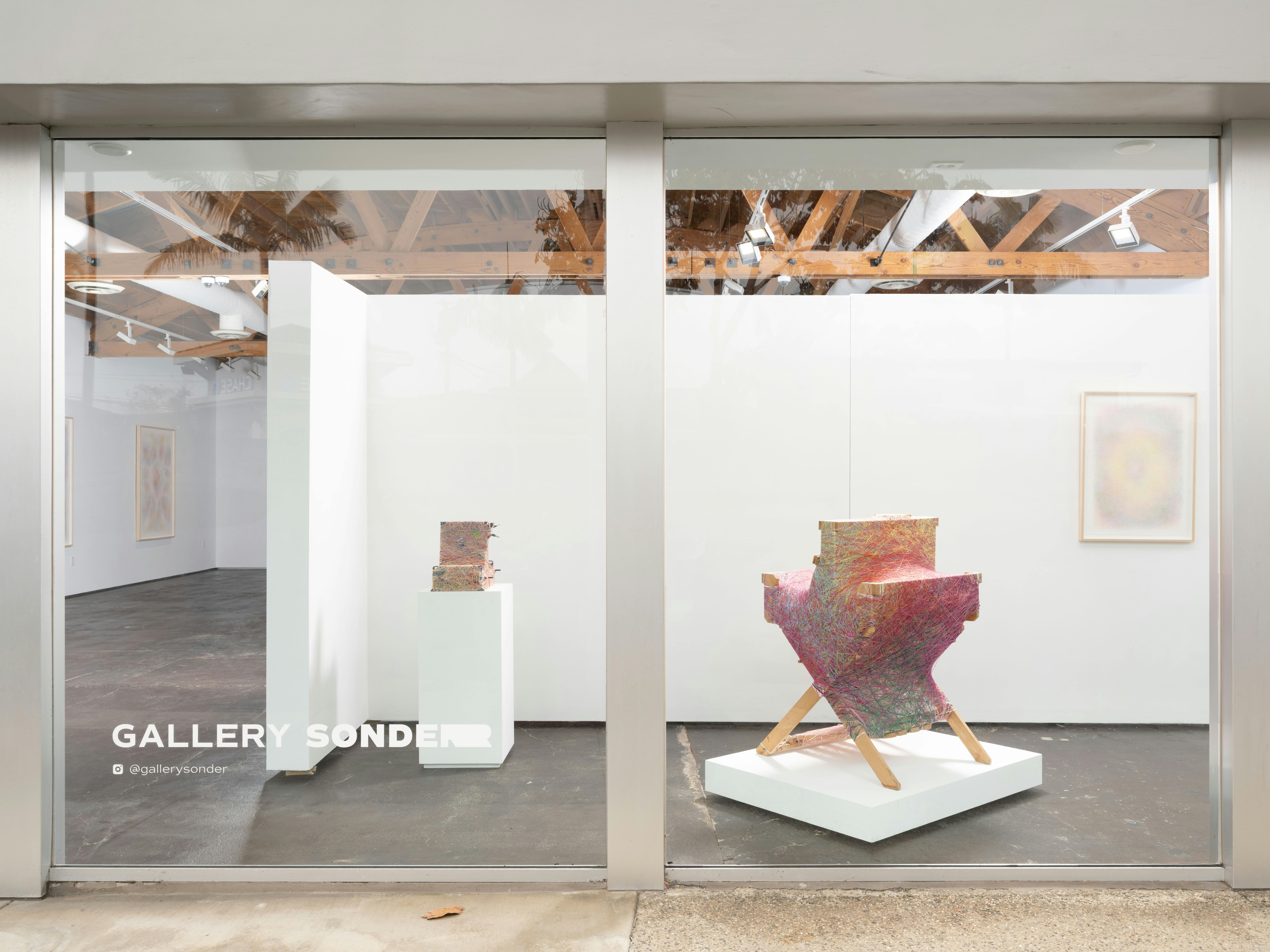 Window-facing installation view of Interstitial, a solo exhibition by Sheng Lor at Gallery Sonder, featuring a colorful sculptural work on a white plinth visible through the gallery facade.