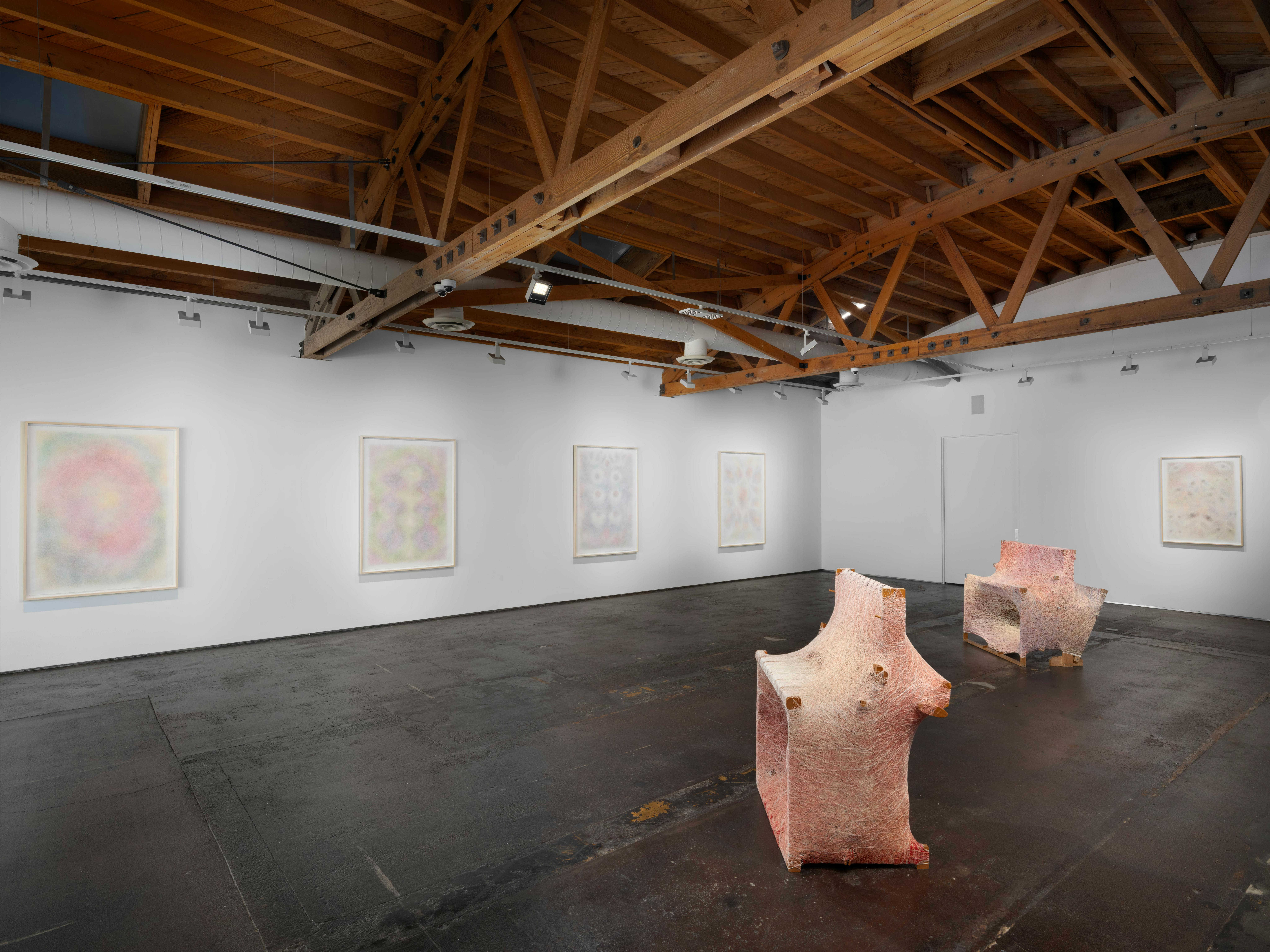 Wide view of the gallery installation showing framed works along the wall with sculptural forms positioned in the foreground.