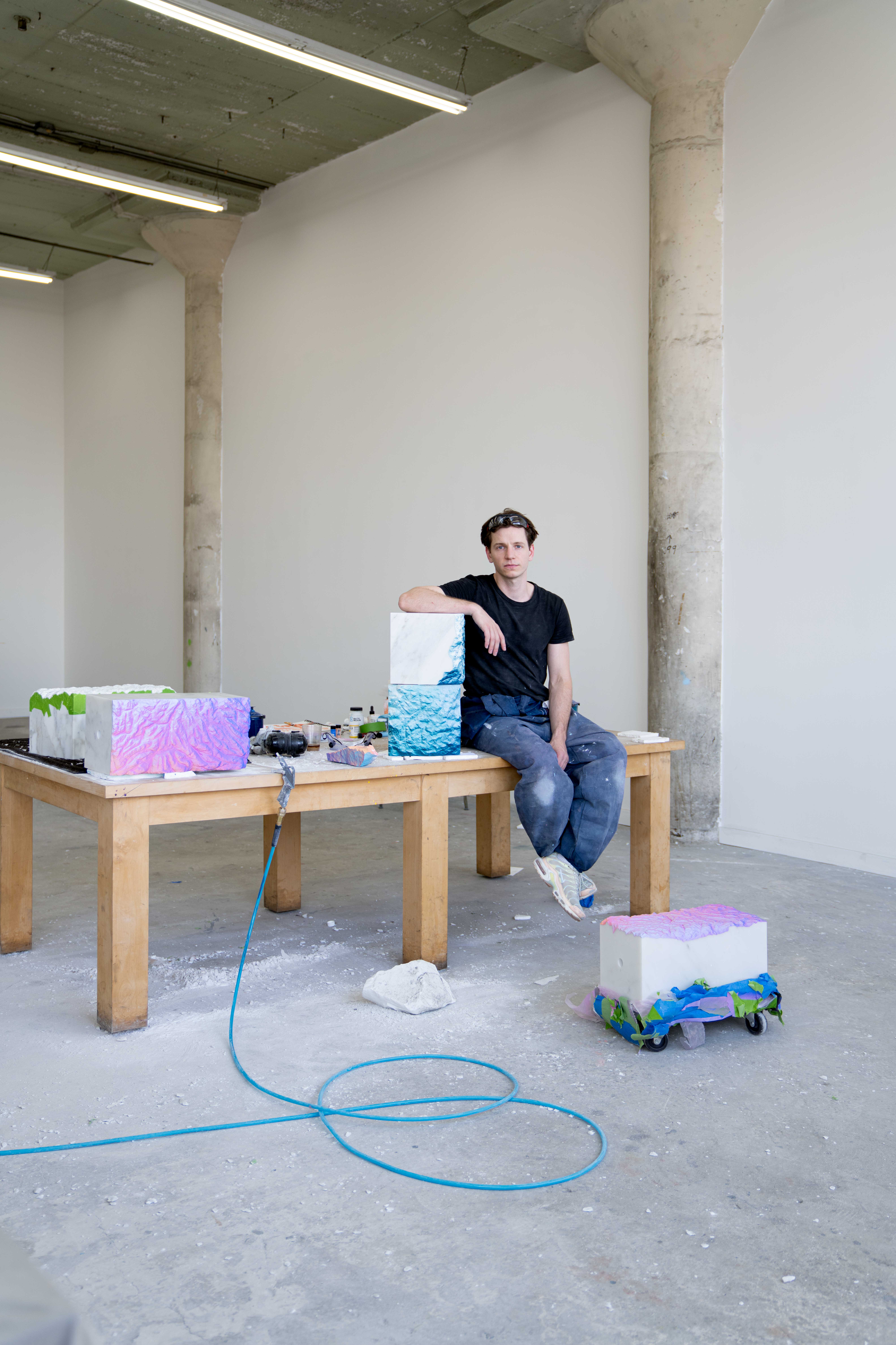 Artist Ruben Benjamin in his studio at Mohlief Studio in Los Angeles, surrounded by works from his Fracture Series, seated among sculptural forms and materials on a worktable, emphasizing process, material experimentation, and studio environment.