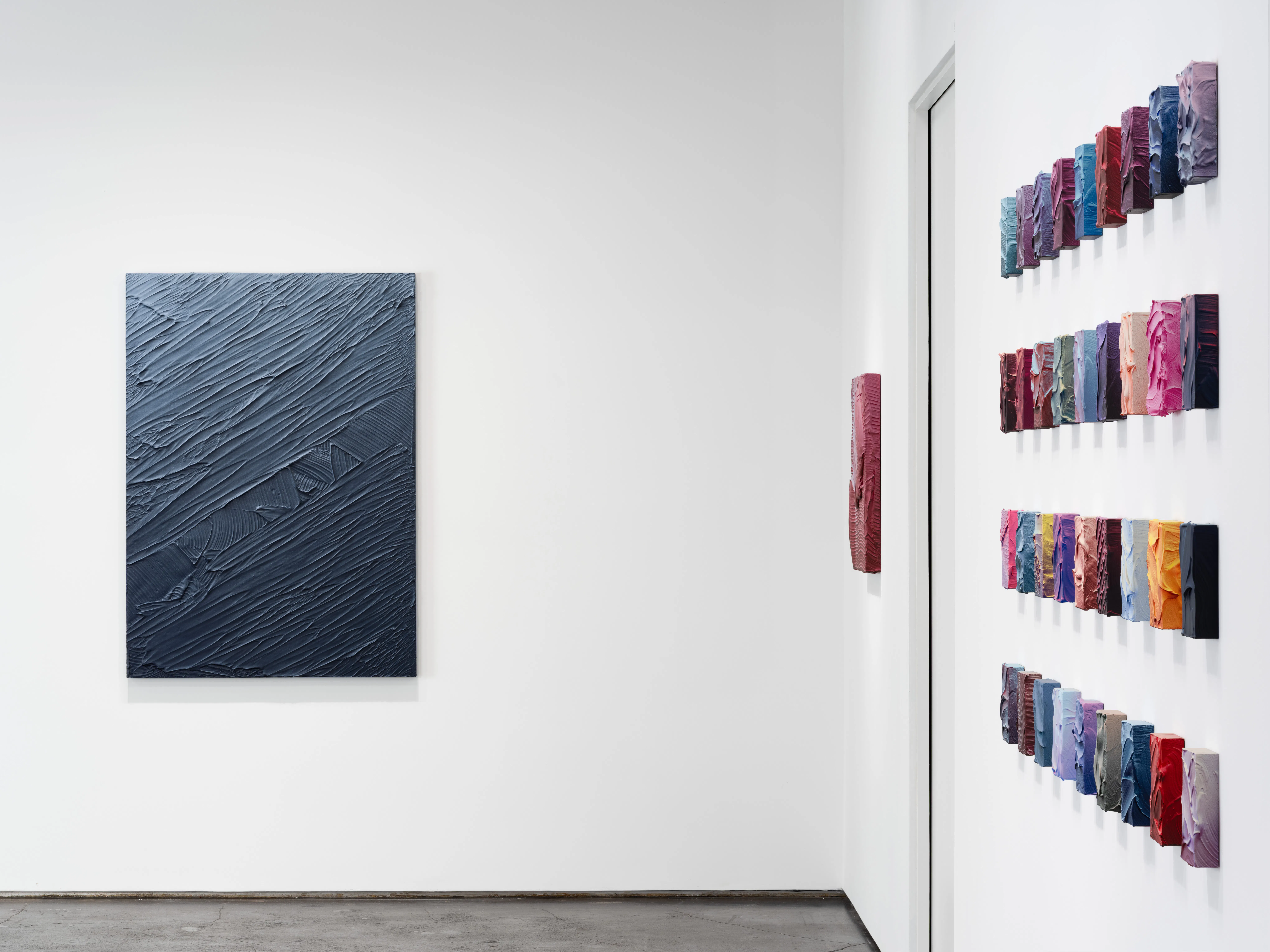 Ruben Benjamin, After The Dust Settles, 2026 — installation view at Gallery Sonder showing a large monochromatic blue impasto painting displayed on a white wall alongside a grid of smaller, vividly colored works arranged in rows, emphasizing contrast, scale, and material variation within the gallery space. Gallery Sonder, Orange County.